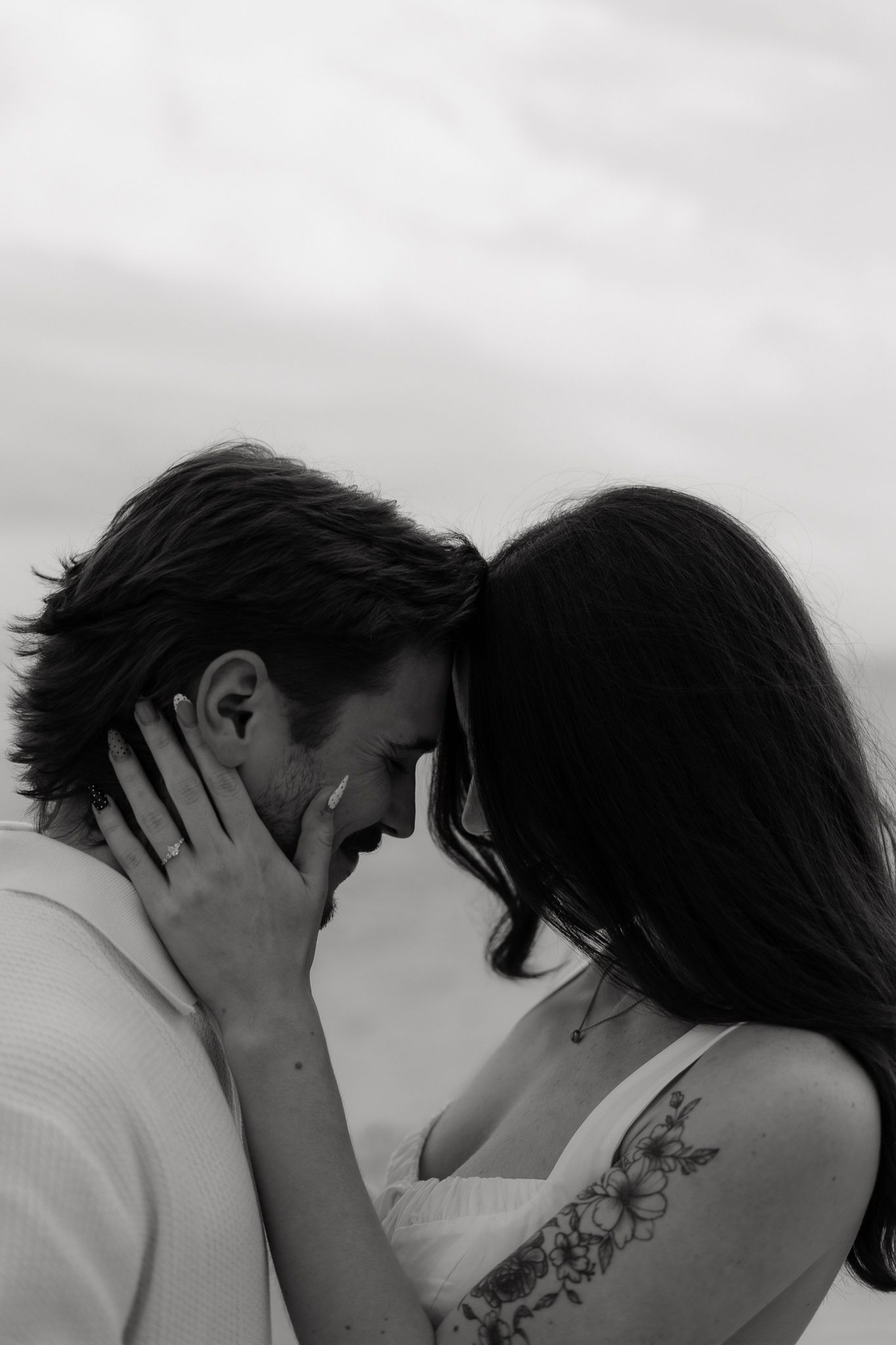 Romantic black and white photo of couple touching foreheads, woman with floral shoulder tattoo embracing man outdoors.