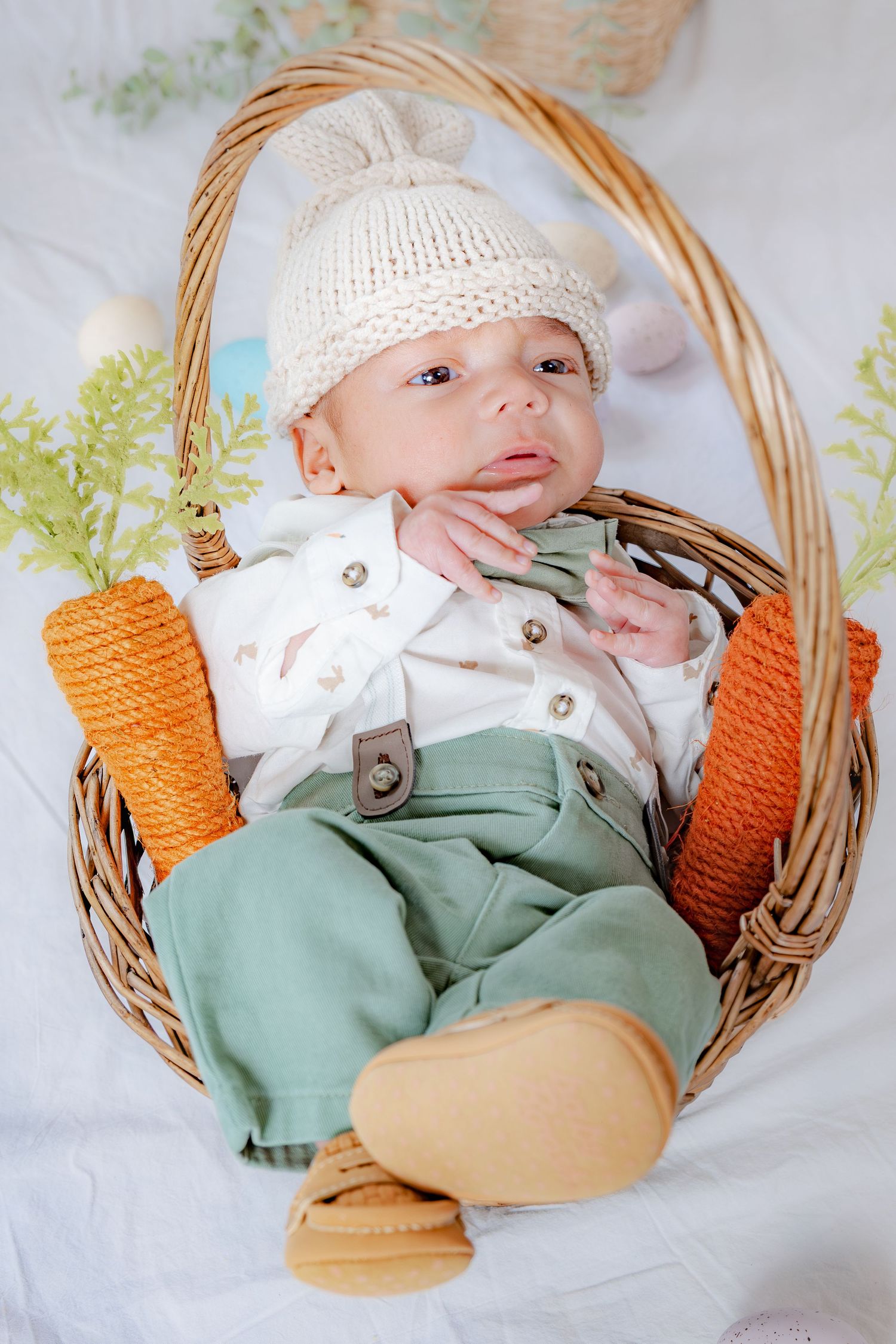 Newborn baby in knit bunny hat sitting in wicker Easter basket with carrot decorations.