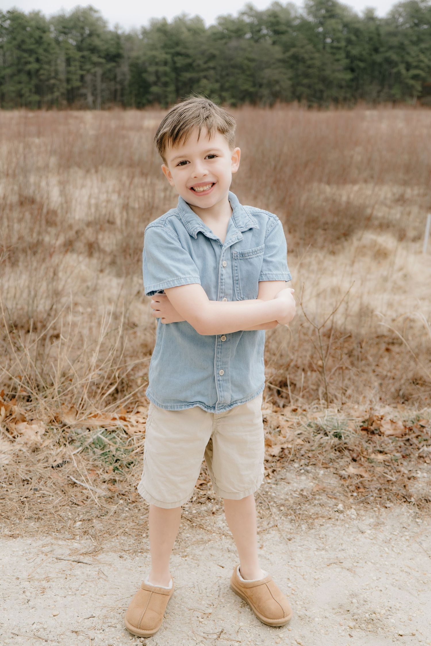 Smiling boy in blue shirt and khaki shorts stands confidently with arms crossed in an open field.
