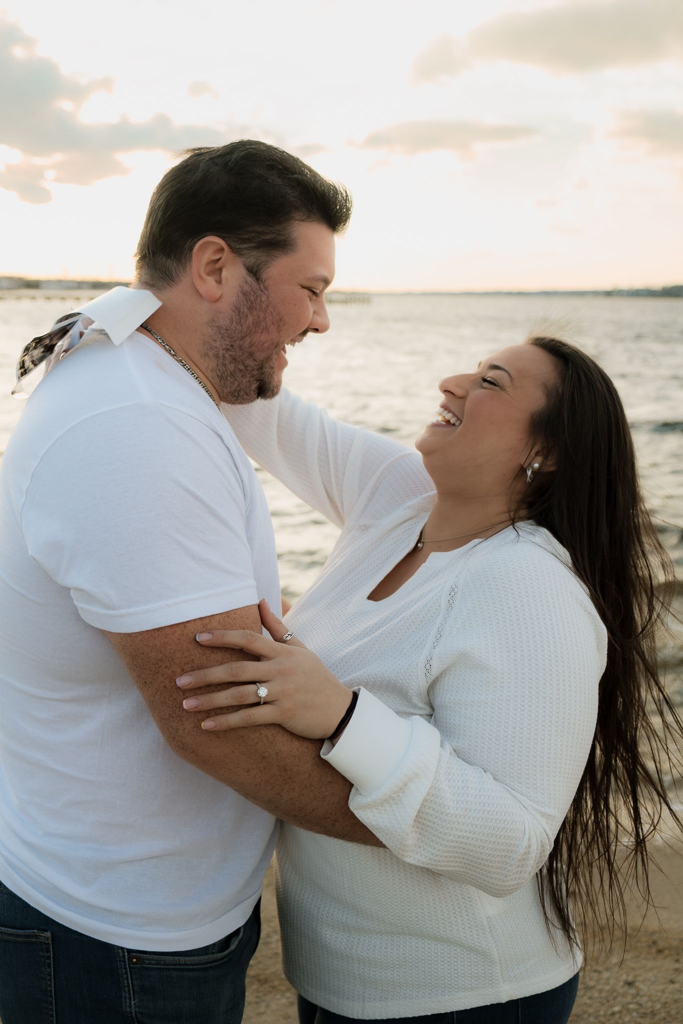 Couple in white tops laughing and embracing on a beach at sunset, woman wearing an engagement ring.
