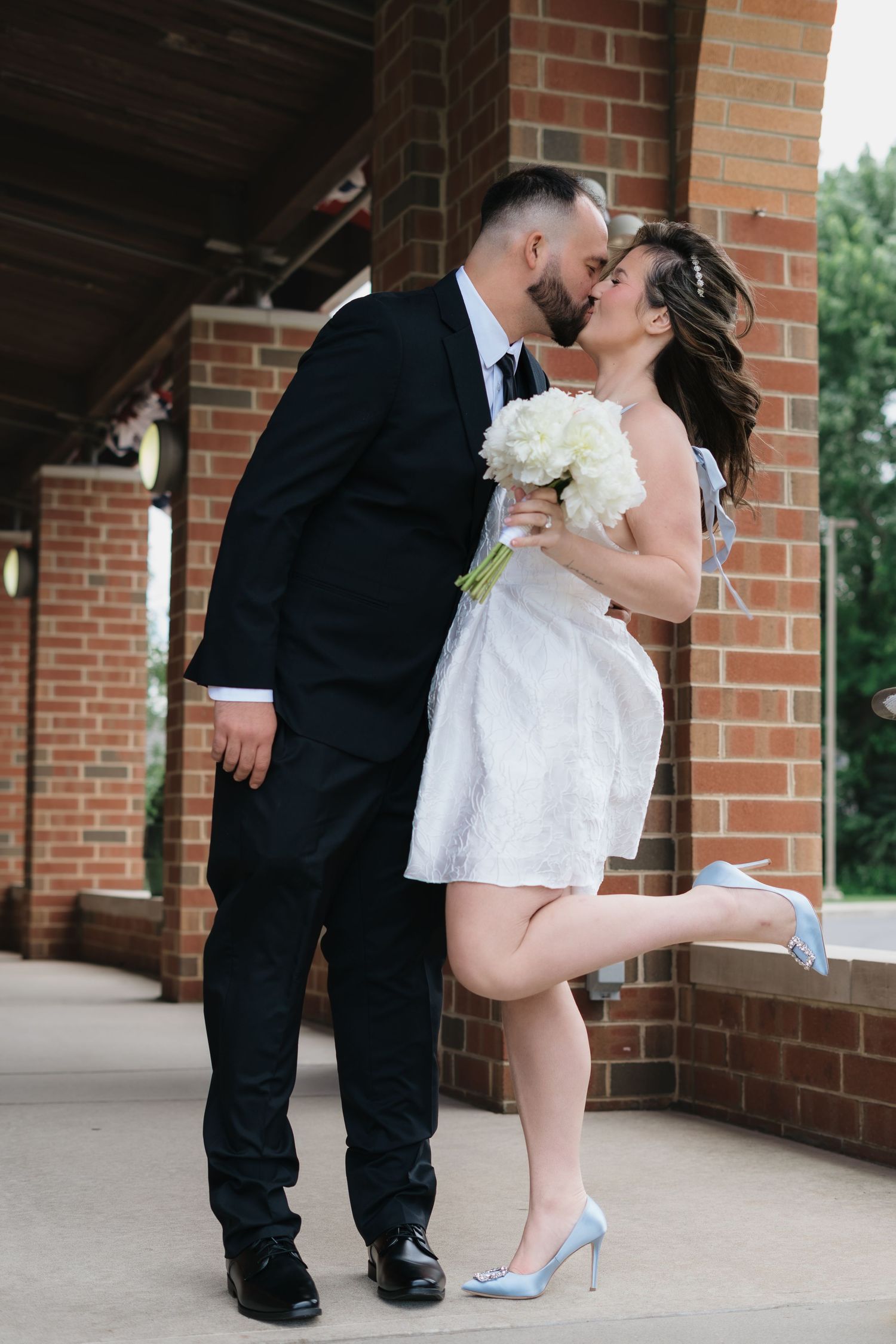 Bride and groom share a romantic kiss outside a brick building, bride holding white bouquet wearing short dress and blue heels.