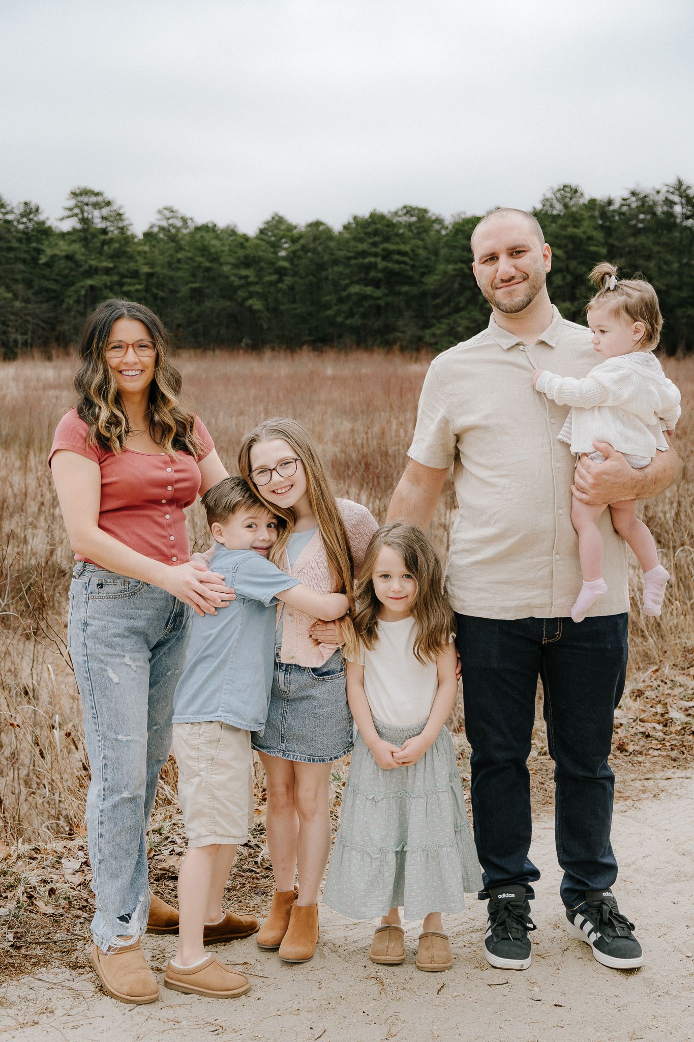 A family of six poses outdoors on a sandy path surrounded by tall grass and pine trees.