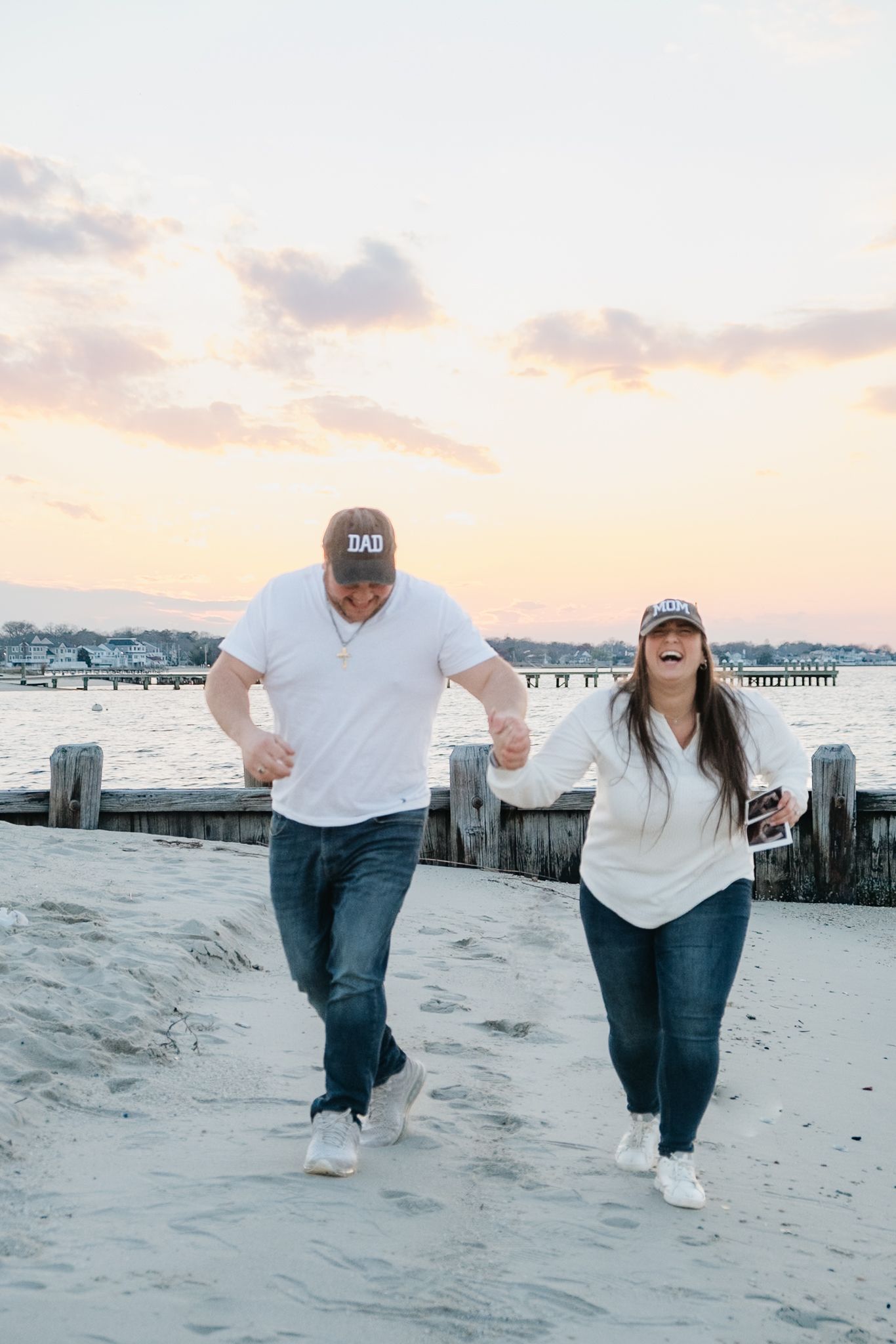 A couple wearing white shirts and dad hats walk hand-in-hand on a sandy beach at sunset near a wooden pier.
