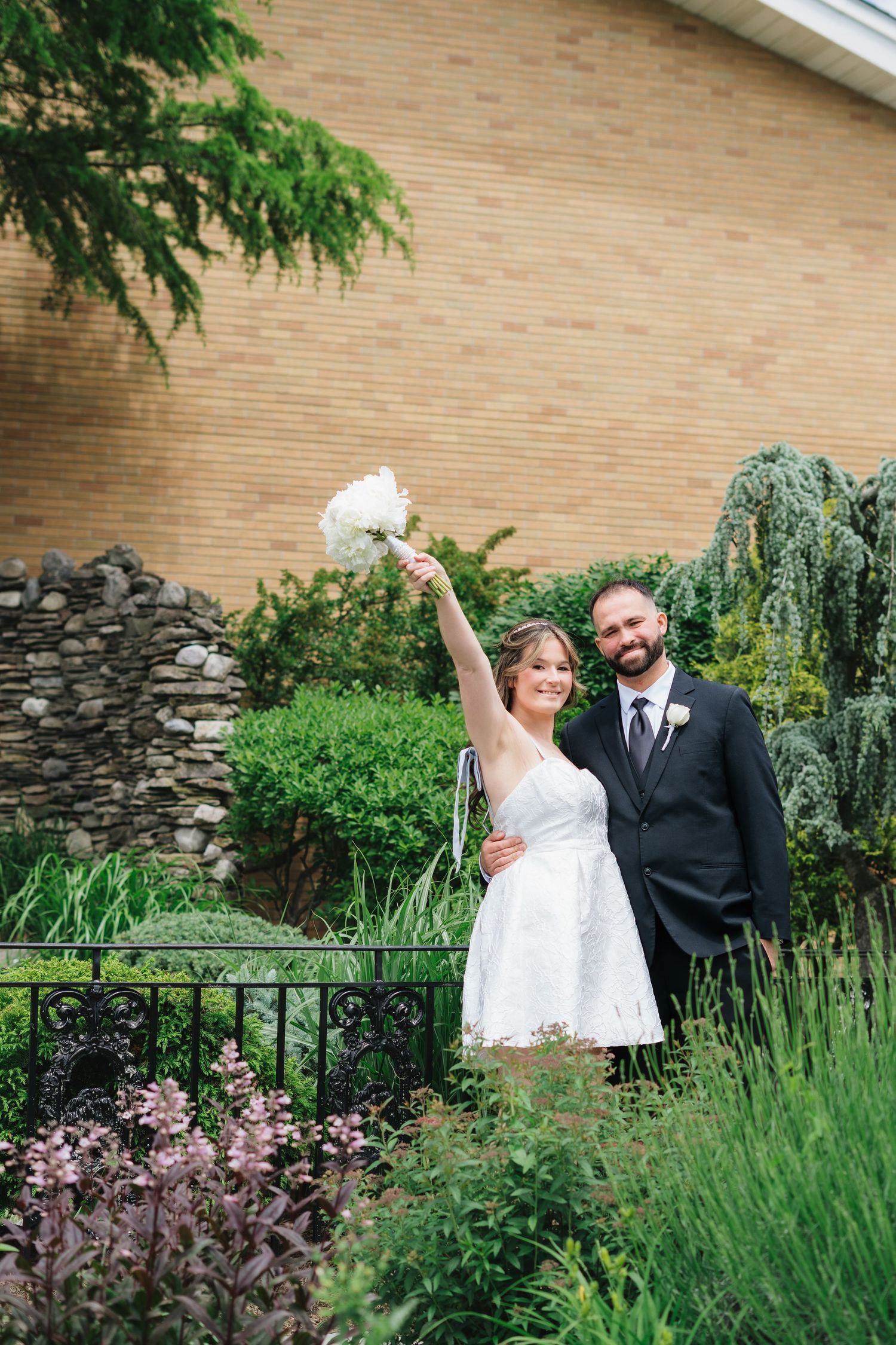 Smiling newlyweds pose in garden, bride raises white bouquet in white mini dress, groom in black suit.