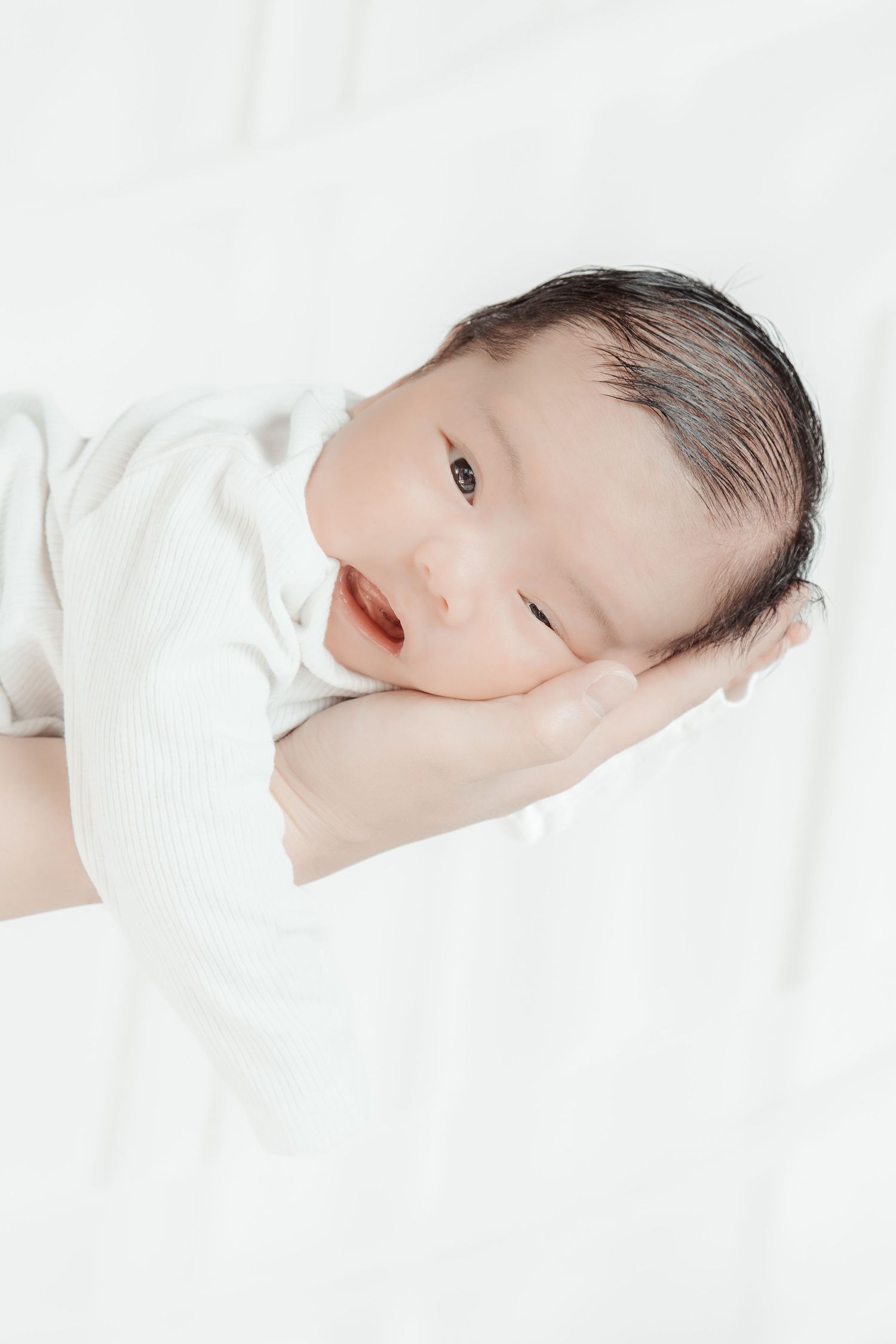 Newborn baby in white onesie resting on a parent's hand against a soft white background.