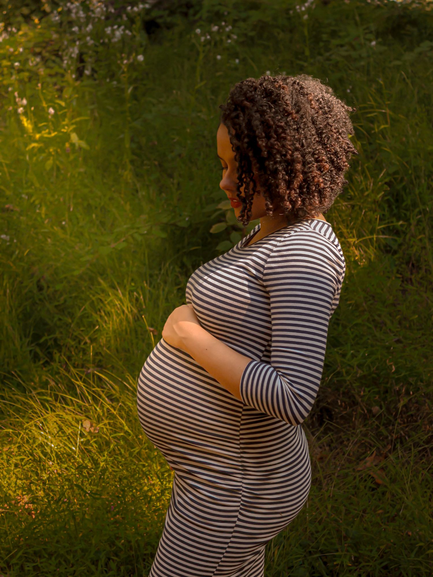 Beautiful pregnant woman in striped dress cradling baby bump while standing in lush green field at golden hour.