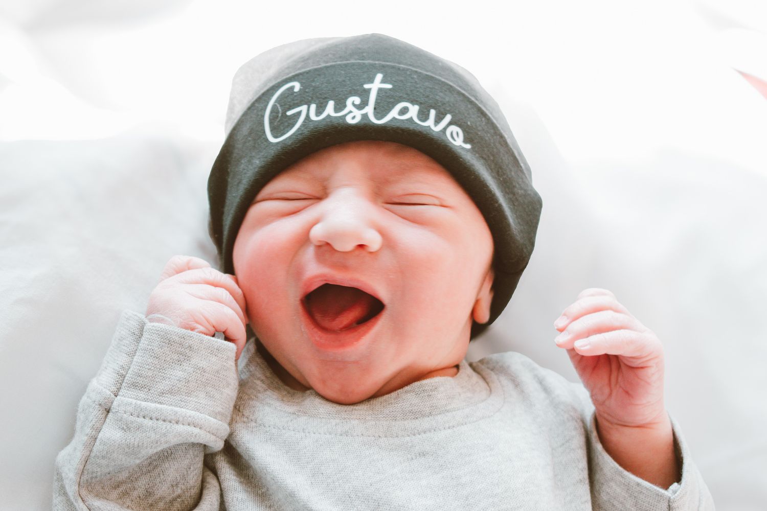 Newborn baby yawning wearing a dark gray beanie hat embroidered with the name Gustavo.