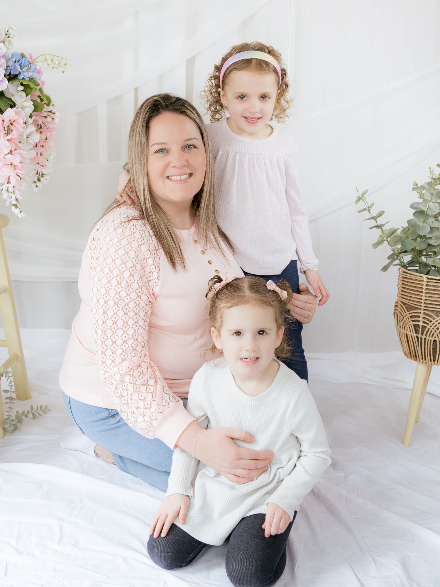 A pregnant mother kneels with her two daughters in coordinating pink outfits in a bright white studio.