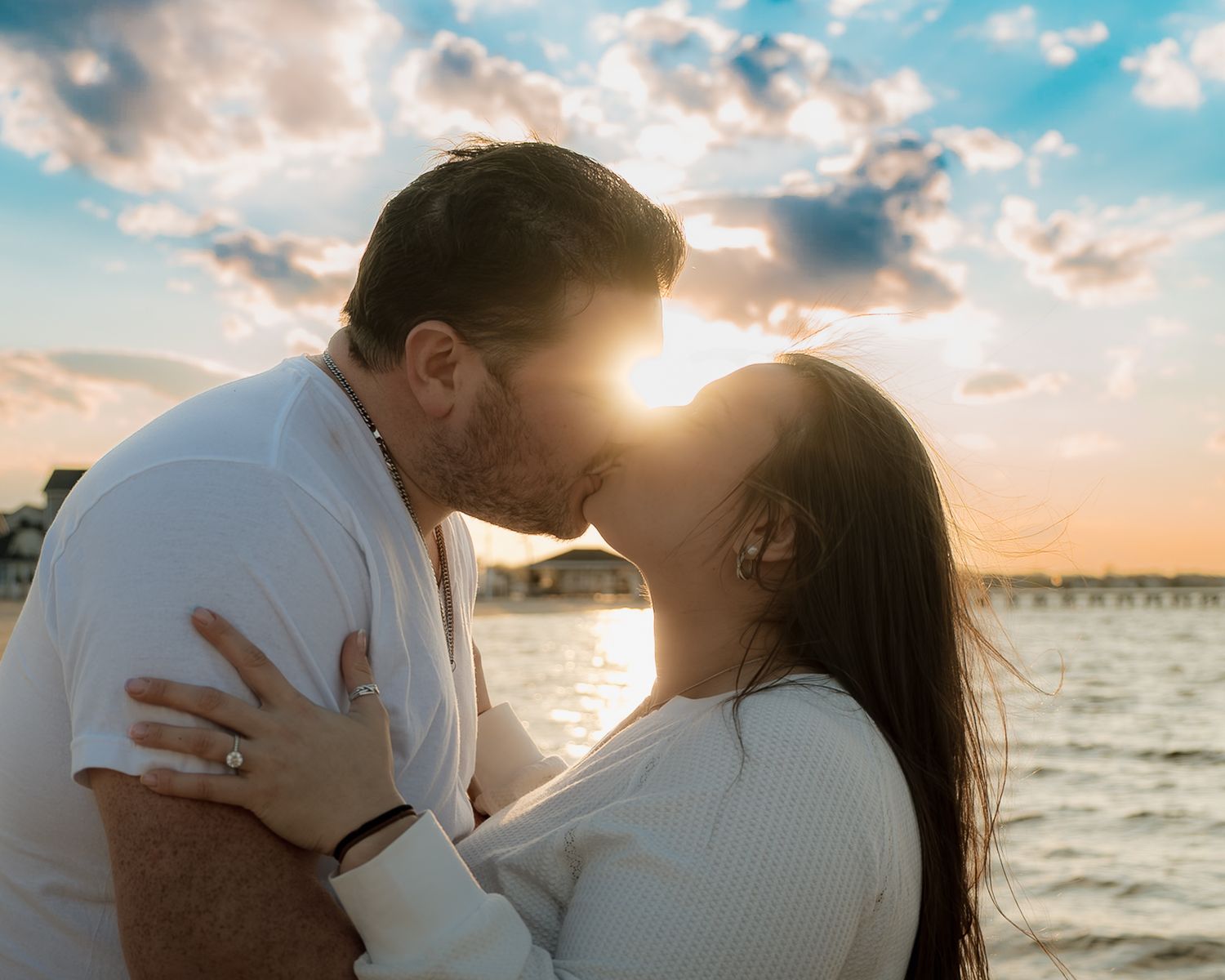 Couple sharing a romantic kiss at sunset by the waterfront with dramatic clouds and golden light behind them.