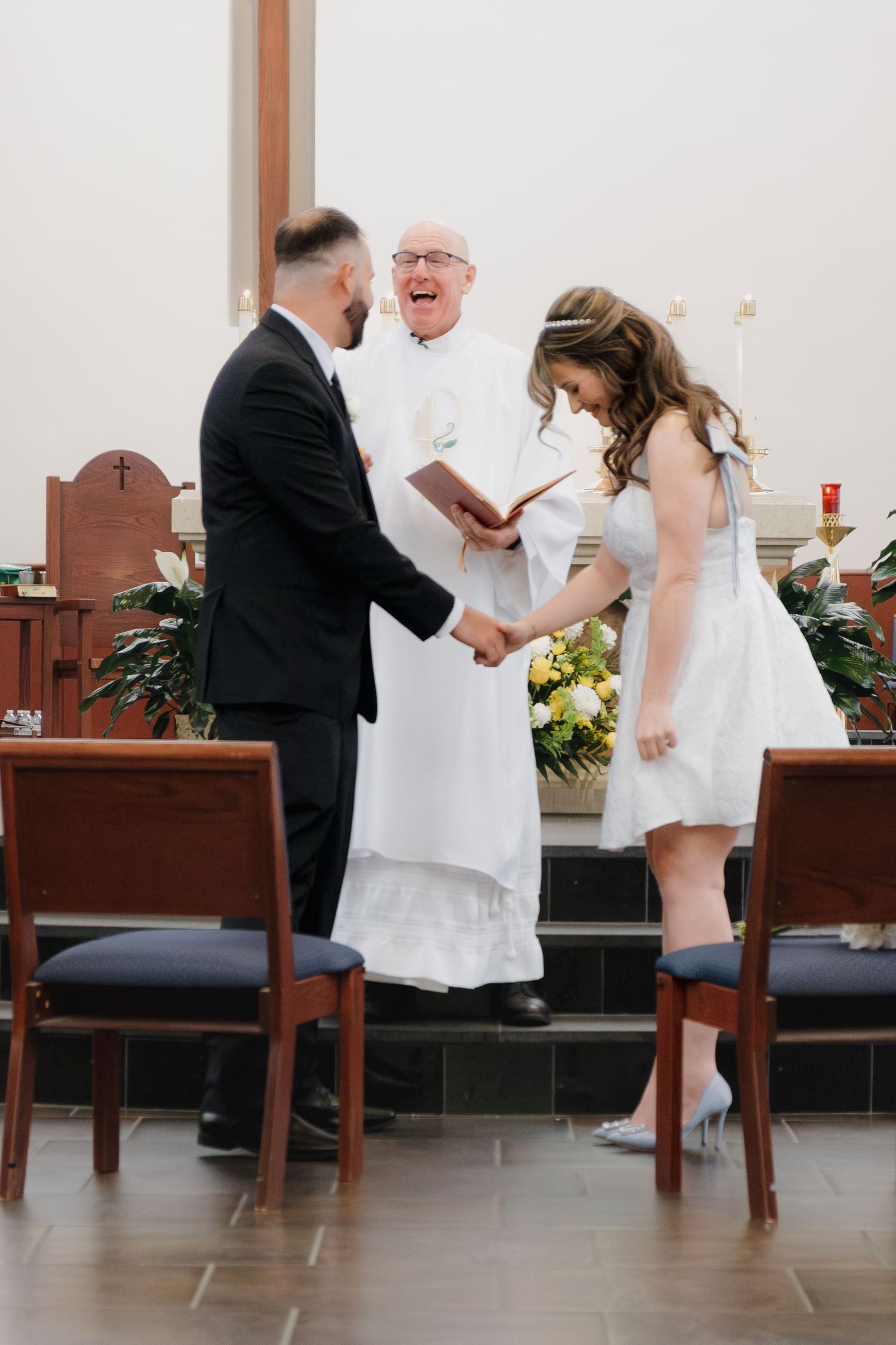 Bride and groom hold hands and laugh with officiant during joyful wedding ceremony at church altar.