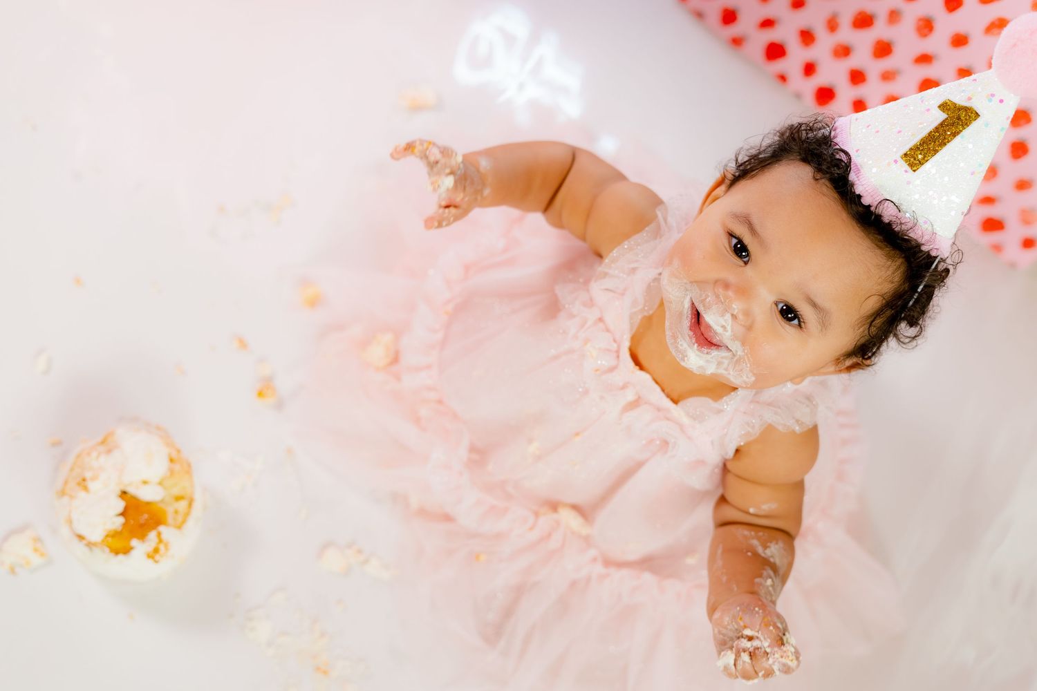 Baby girl in pink tutu dress with cake smash on face wearing birthday hat, celebrating first birthday.