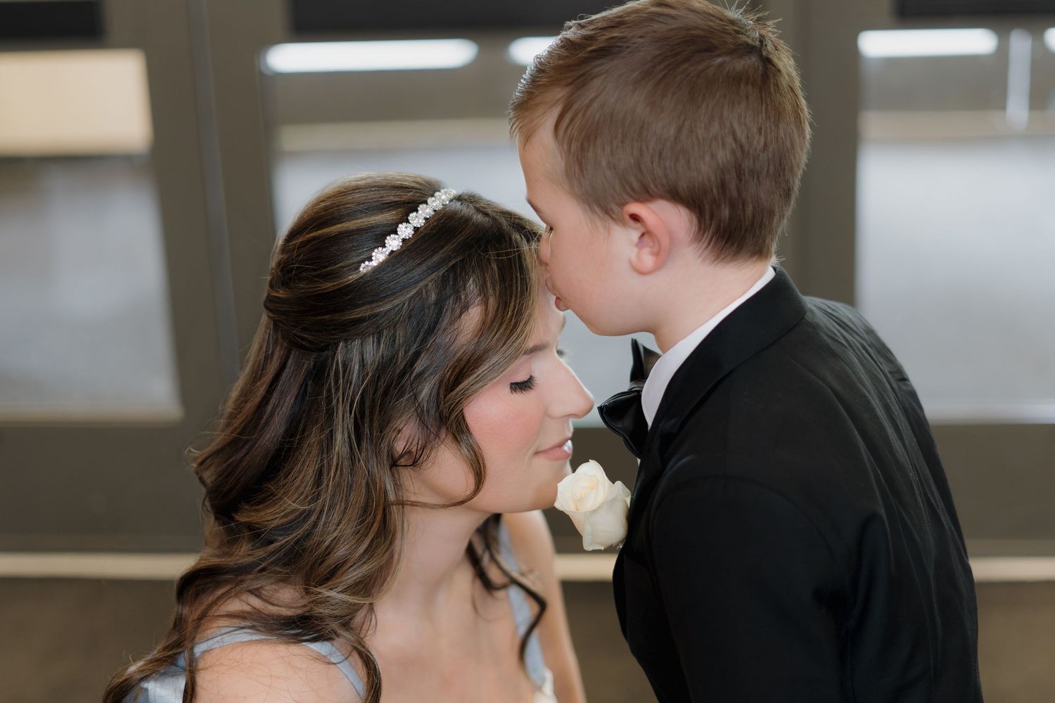Bride in white wedding dress shares sweet moment with young boy in black tuxedo at wedding celebration.