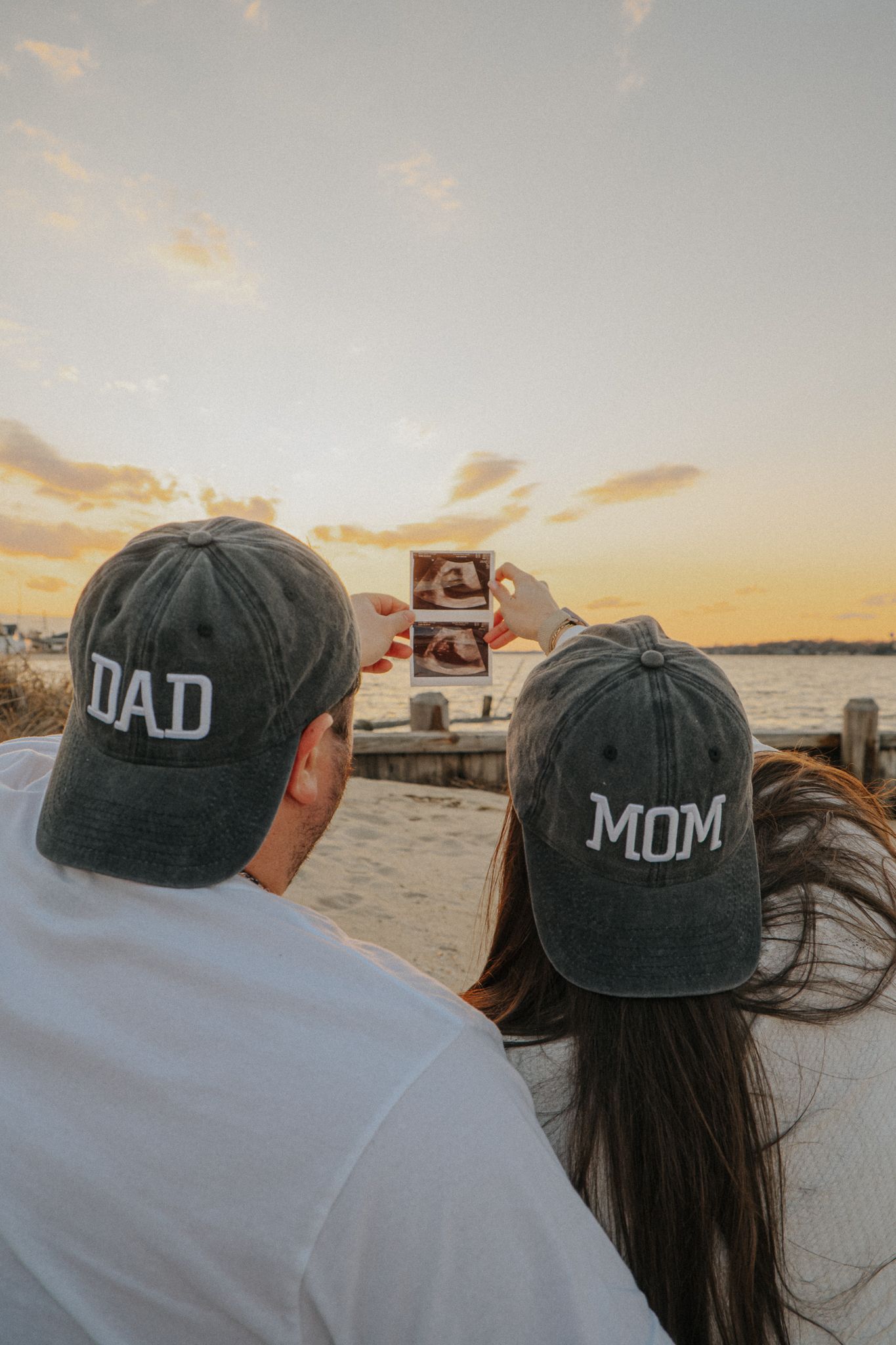 Couple wearing Dad and Mom hats holding ultrasound photo at sunset by the water, pregnancy announcement.