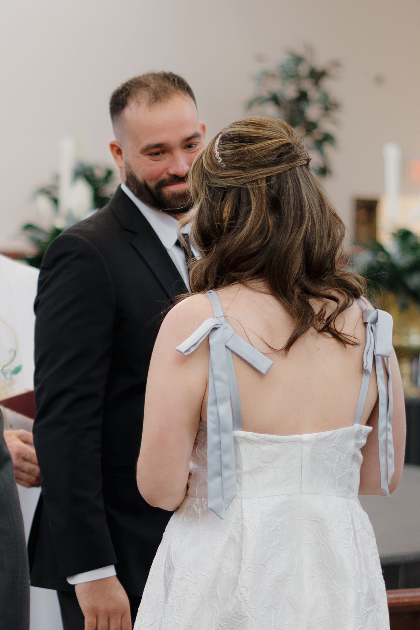 Groom in black suit smiling at bride in white dress with blue ribbon ties during wedding ceremony.