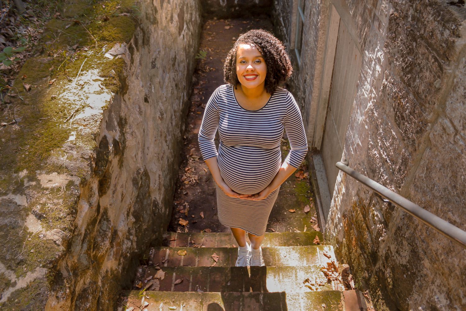 Happy pregnant woman in striped maternity dress standing at bottom of rustic stone stairway during outdoor photoshoot.