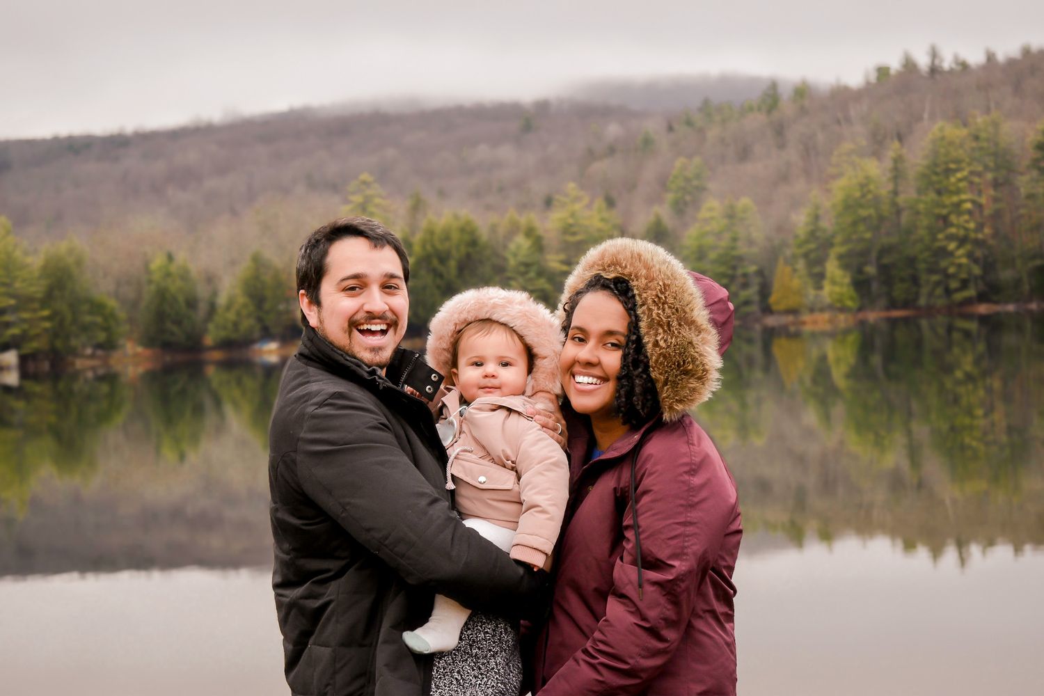 Happy family with baby posing by a lake with forested hills in the background on a cloudy day.