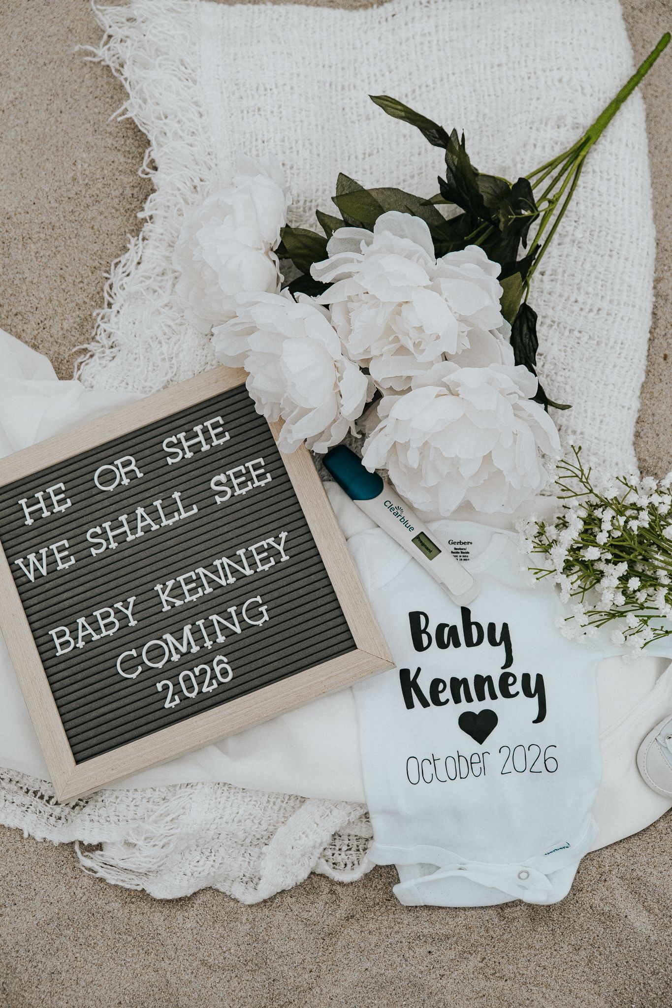 Baby Kenney pregnancy announcement with letter board, onesie, and white flowers on sand, due October 2026.
