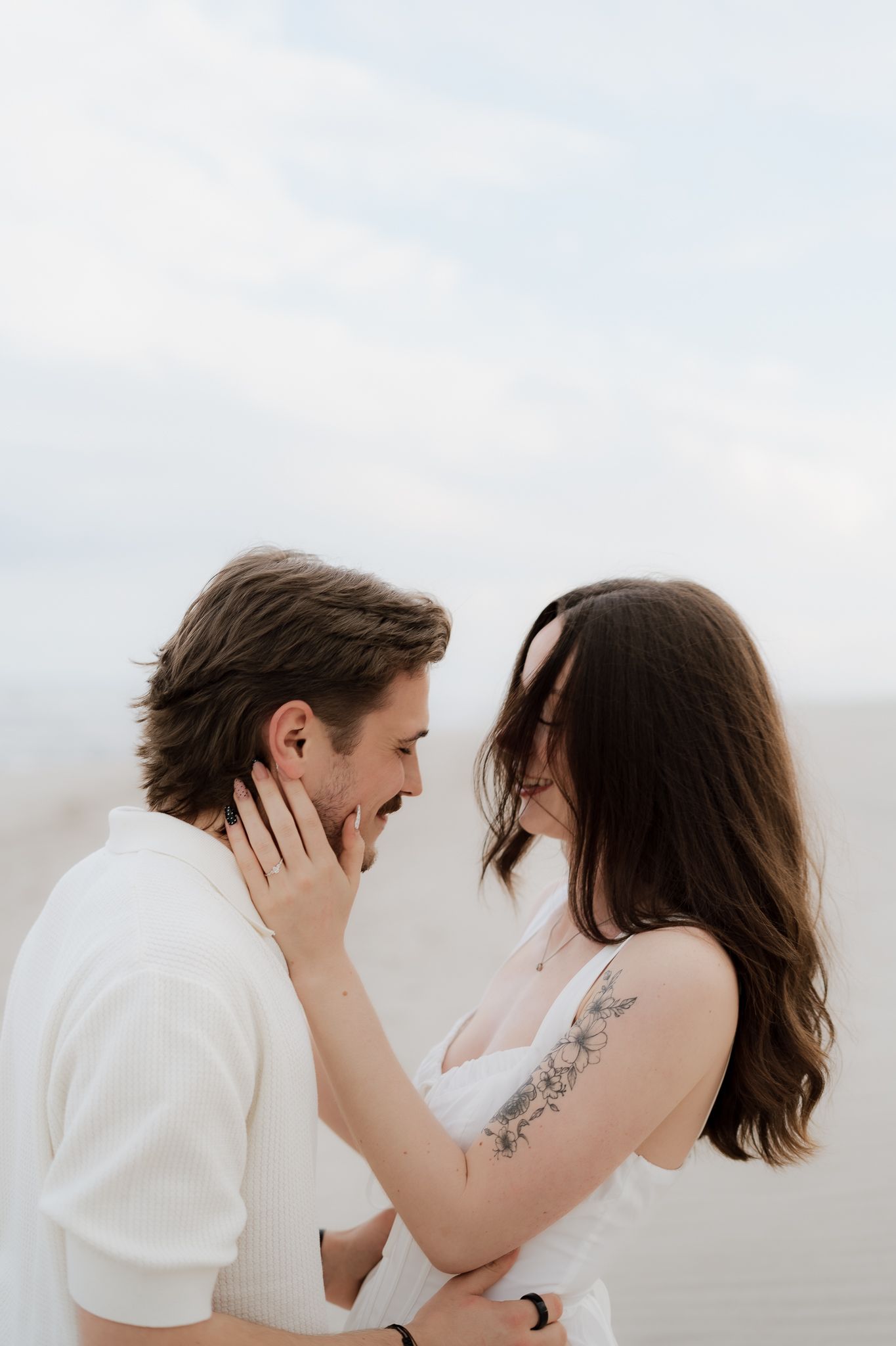 Couple in white outfits sharing a tender moment, woman with floral arm tattoo gently cupping mans face at the beach.