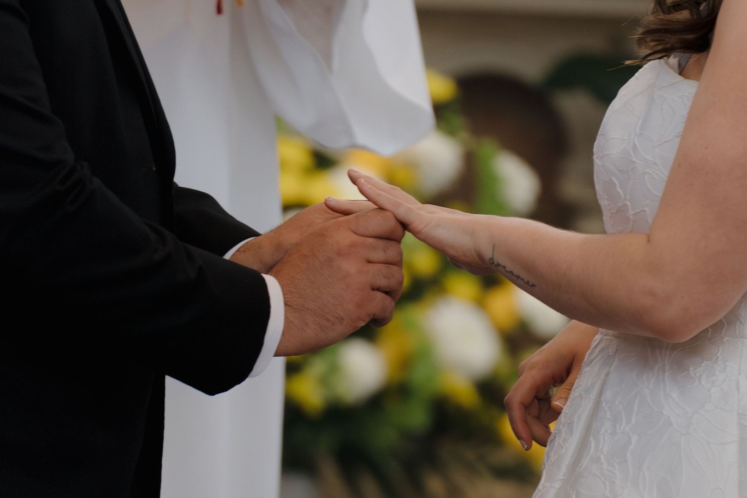 Groom placing a wedding ring on bride's finger during a ceremony with yellow and white flowers in background.