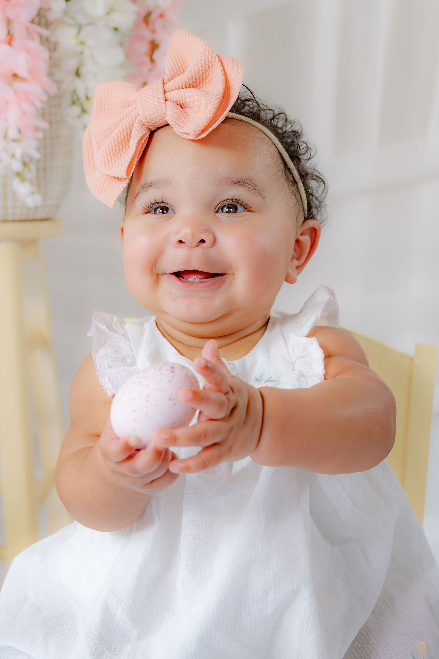 A happy baby girl in a white dress and peach bow headband holds a pink Easter egg while smiling brightly.
