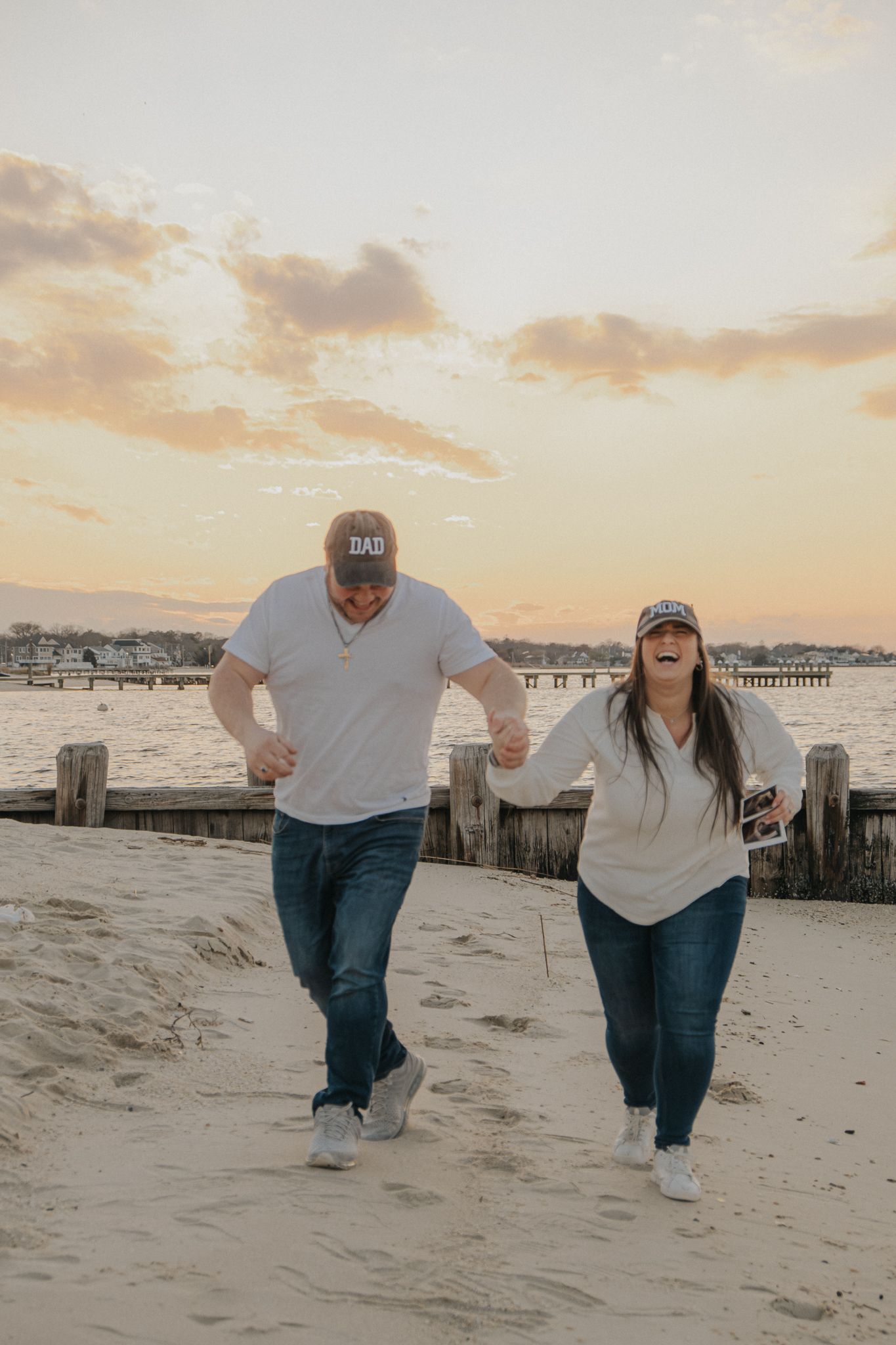 Couple laughing and walking on sandy beach at sunset, wearing matching caps and jeans near wooden pier.
