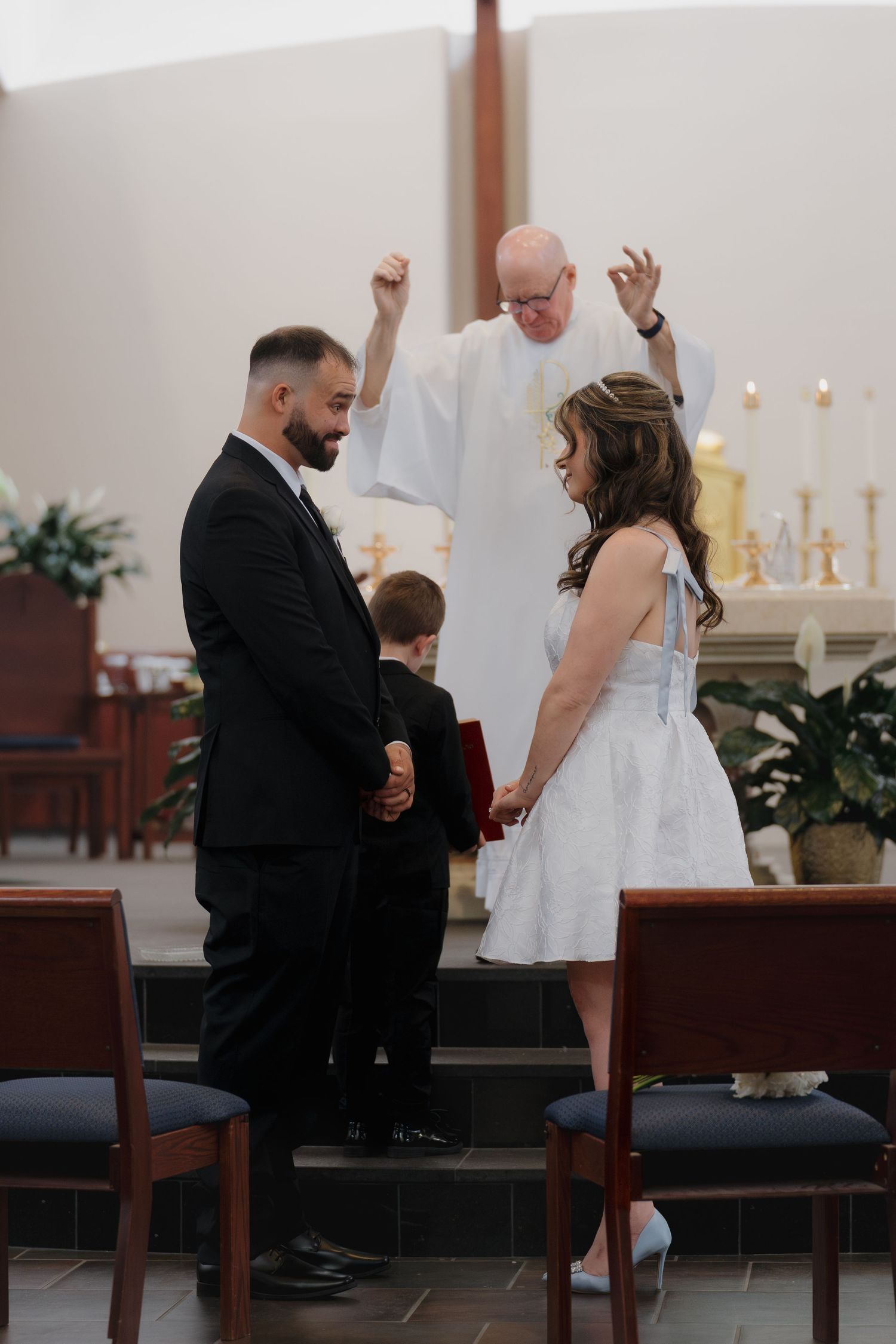 Couple exchanges vows at church altar with priest blessing them, young boy stands beside groom in black suit.