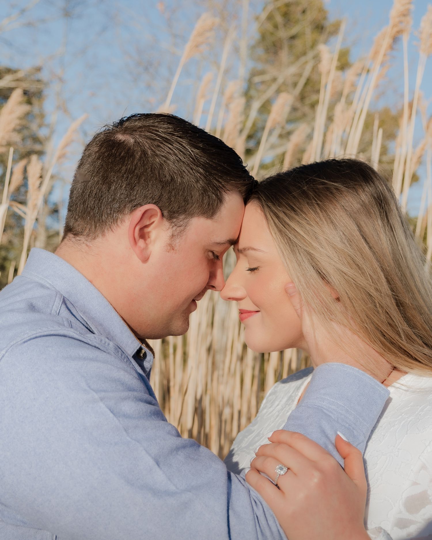 Couple sharing a romantic forehead touch outdoors near tall golden reeds, woman wearing an engagement ring.