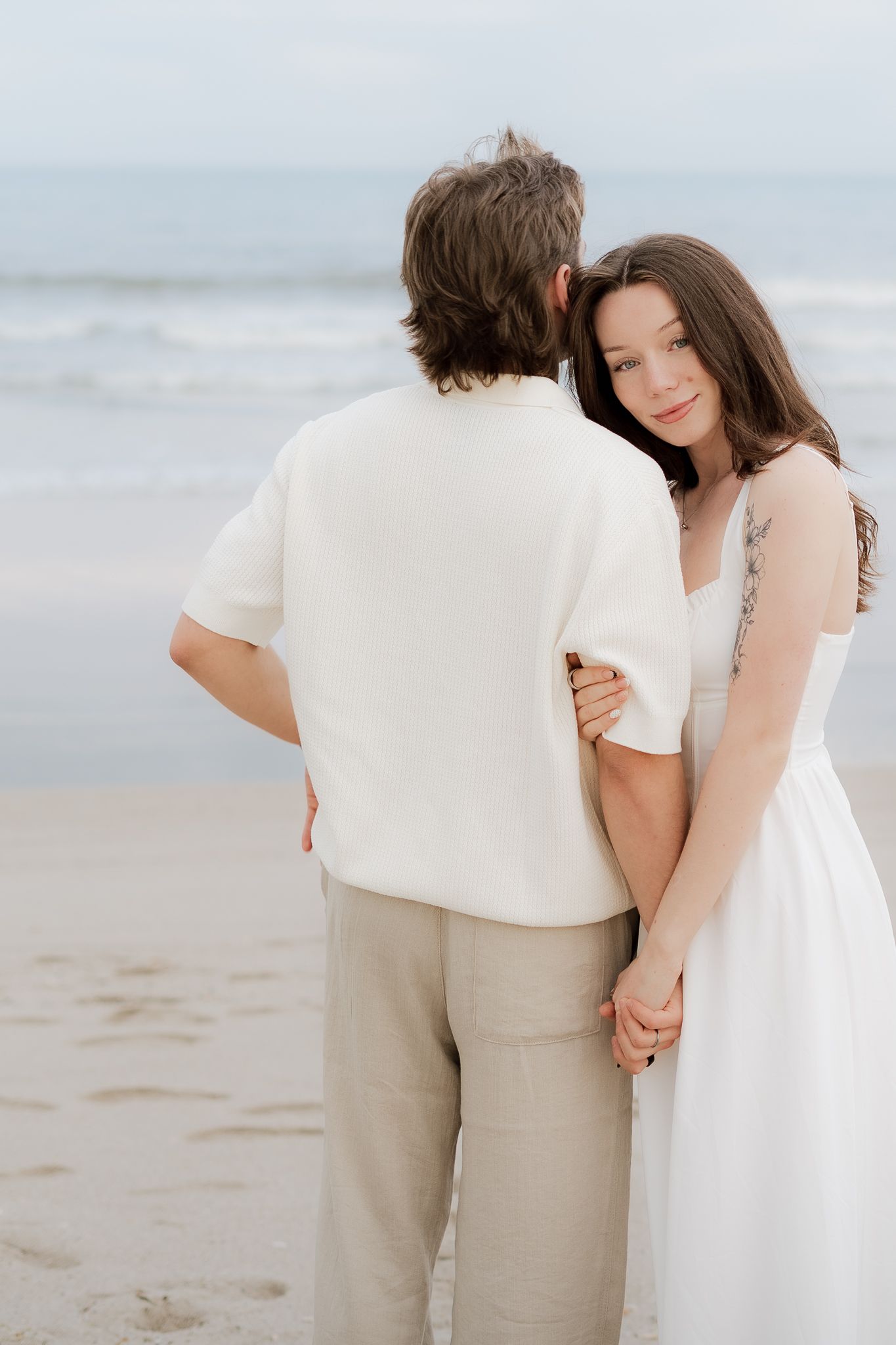 Couple holding hands on a beach, woman in white dress with floral arm tattoo leans on mans shoulder.