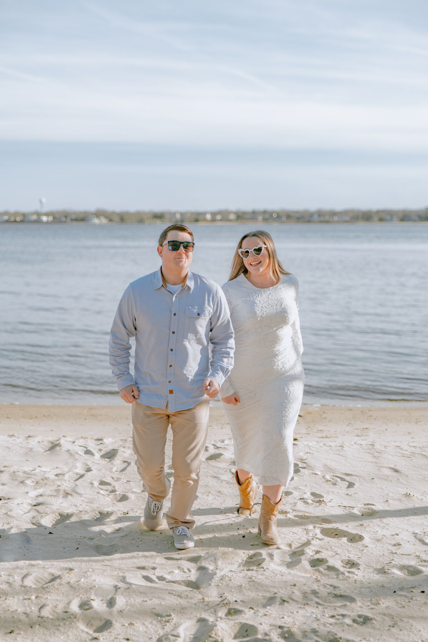 Happy engaged couple walking hand-in-hand on a sunny beach wearing sunglasses and casual attire.