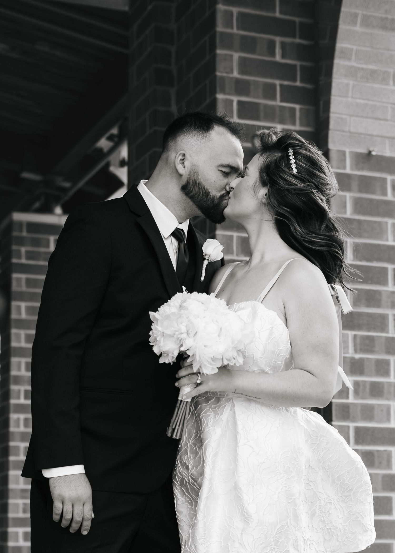 Black and white photo of bride and groom kissing outside brick building, bride holding white floral bouquet.