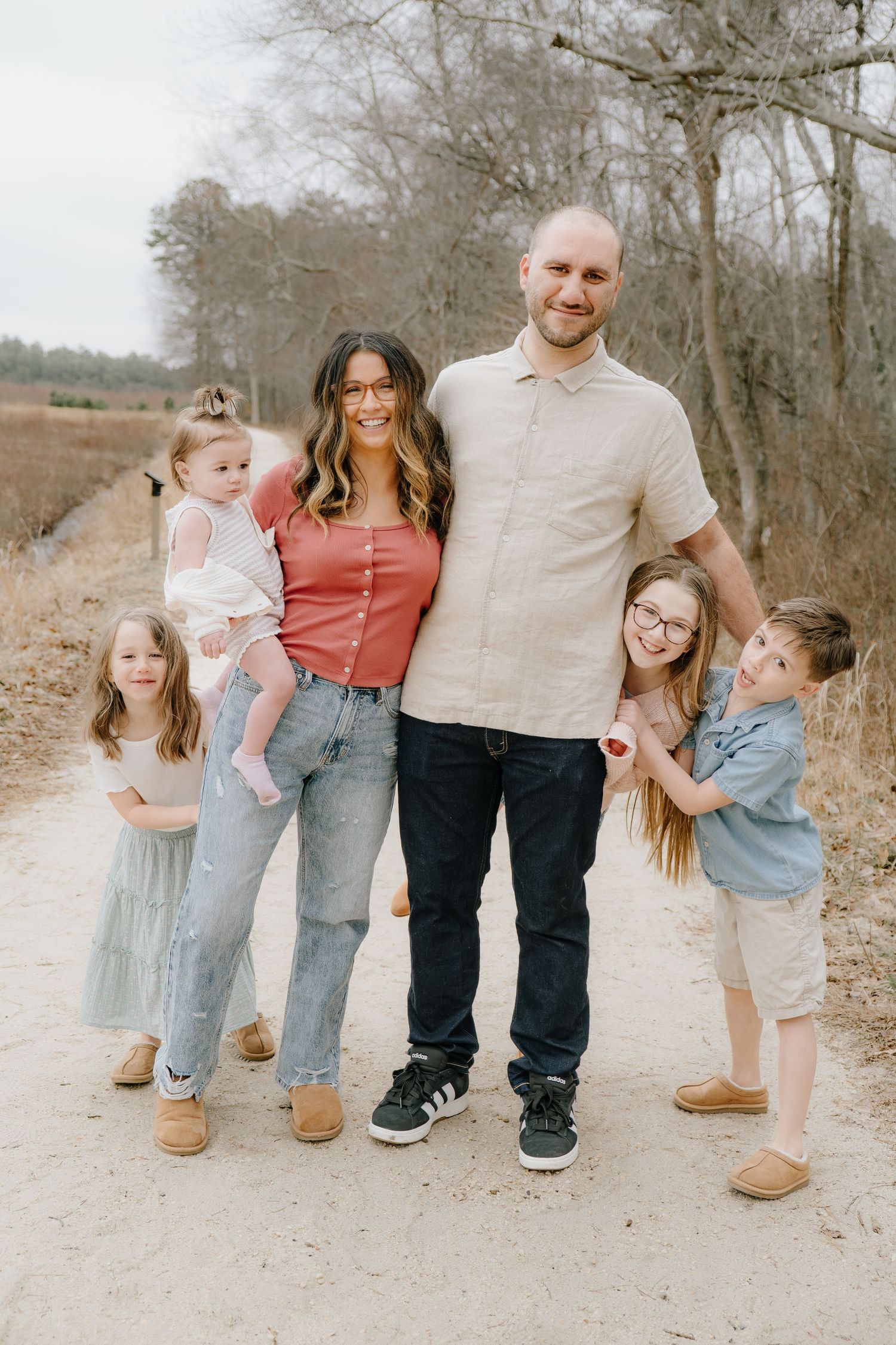 Family of six posing on a dirt path surrounded by bare winter trees, smiling together outdoors.