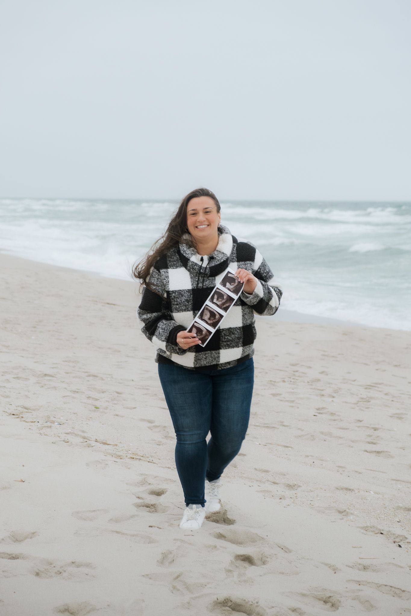 Smiling pregnant woman holding ultrasound photos on a sandy beach wearing a black and white plaid jacket and jeans.