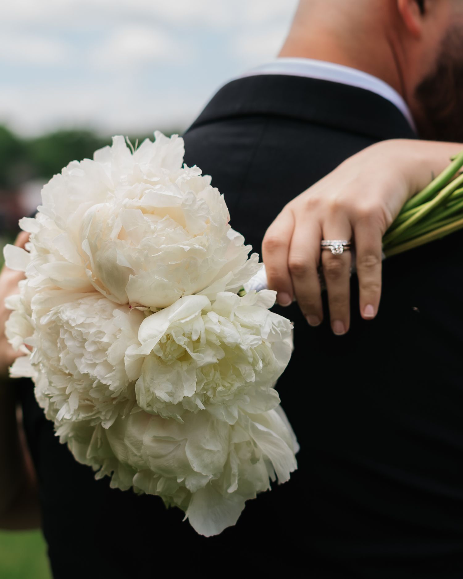 Bride holding white peony wedding bouquet while embracing groom in black suit, diamond engagement ring visible.