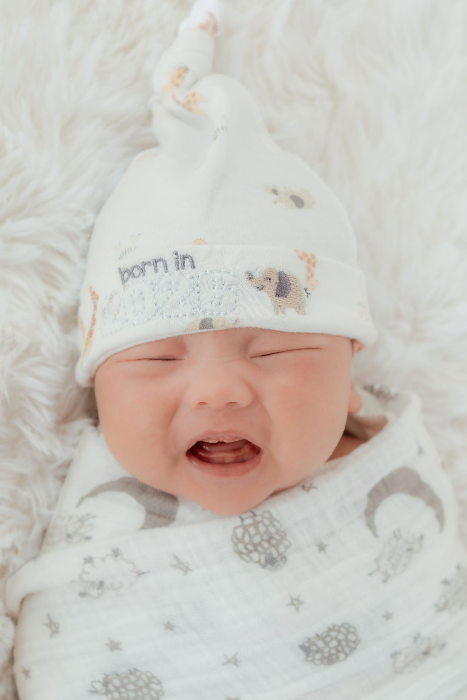 A crying newborn baby wrapped in a white swaddle blanket wearing a white knot hat that reads born in.