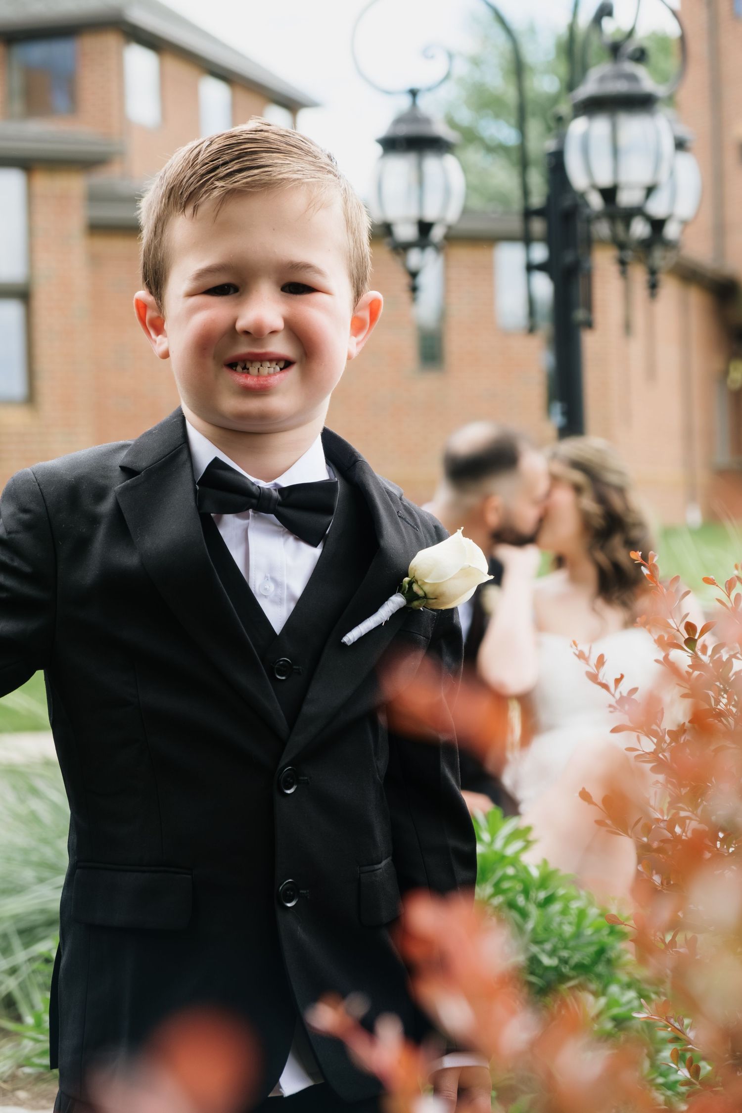 Smiling young boy in black tuxedo with boutonniere poses in foreground while bride and groom kiss behind him.