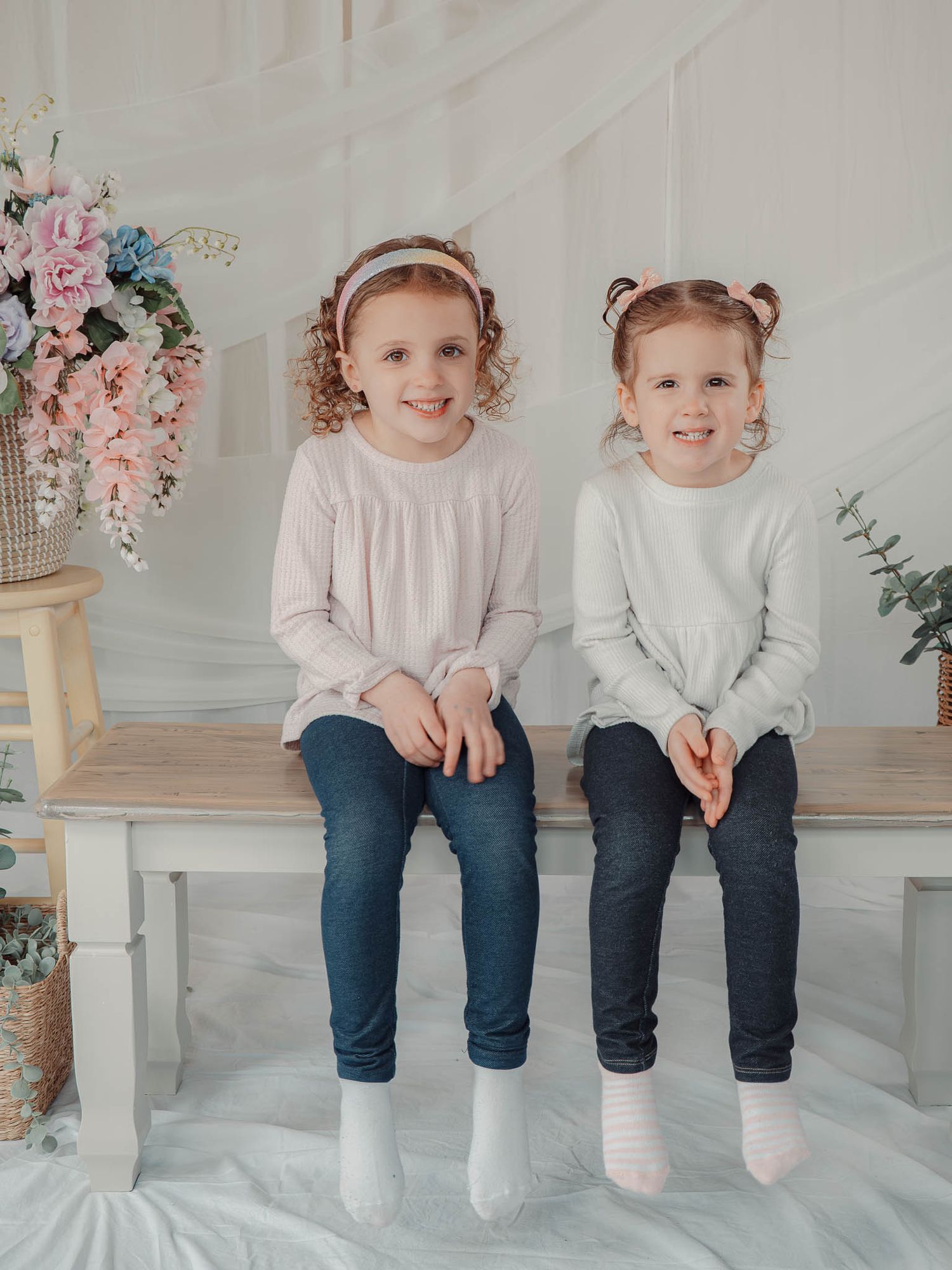 Two young red-haired sisters in pastel tops and jeans sit together on a bench in a bright floral studio.