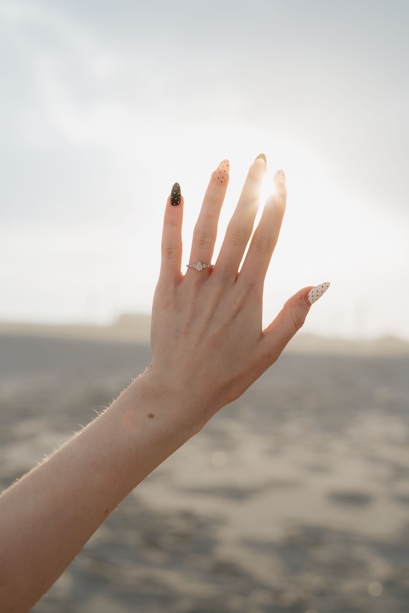 Close-up of a woman's hand displaying an engagement ring on a sunlit sandy beach.