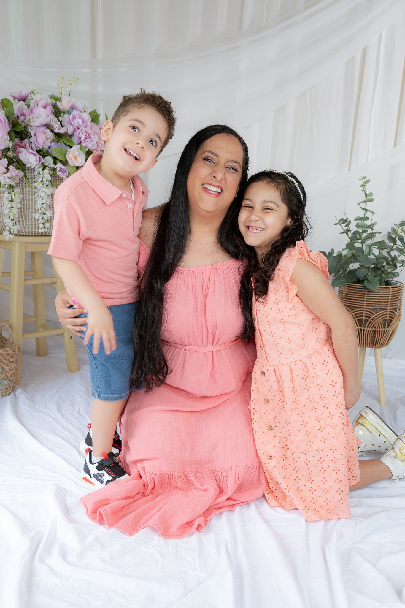A mother in a pink dress sits with her young son and daughter in a bright floral studio.