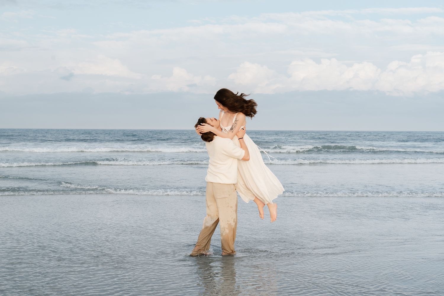 Couple embracing on a beach, man lifting woman in white dress near ocean waves under a cloudy sky.
