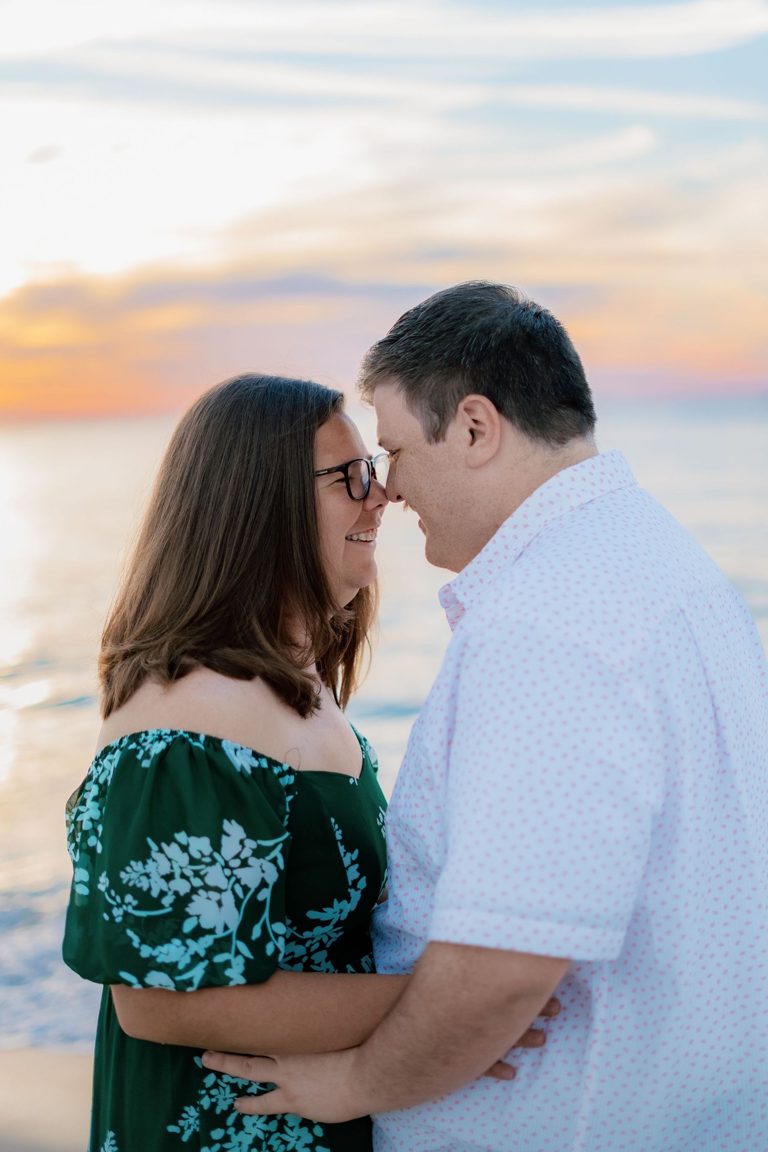 Couple sharing a romantic nose-to-nose moment on the beach at sunset, woman in green floral dress, man in white shirt.