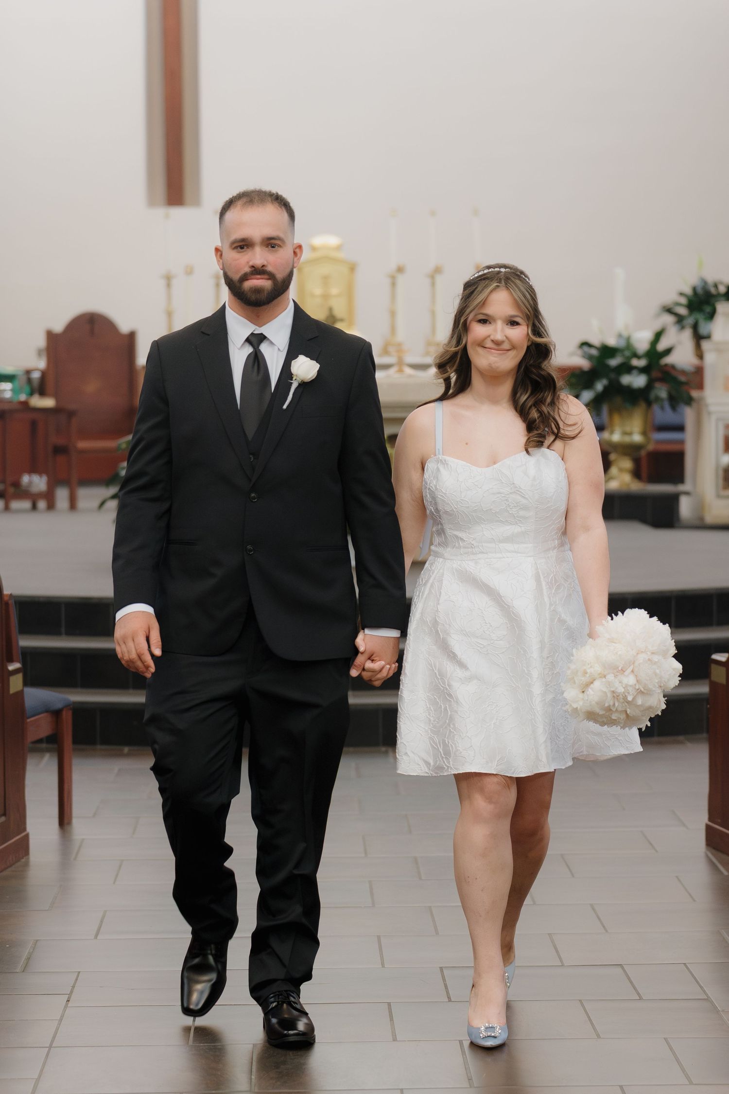 Bride and groom walk hand in hand down church aisle, bride in short white dress holding white peony bouquet.