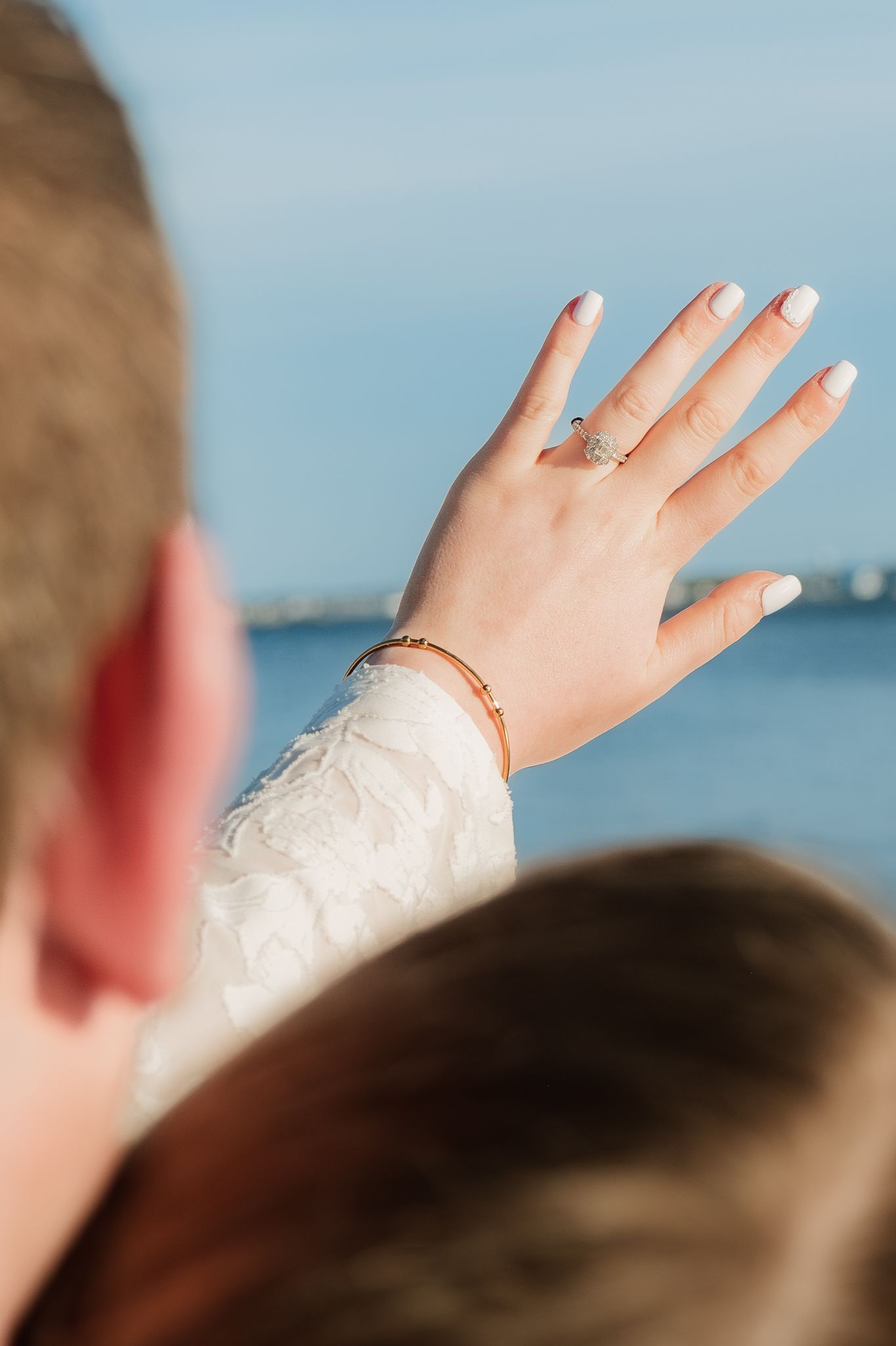 Close-up of a woman's hand displaying an engagement ring and gold bracelet, with a scenic waterfront in the background.