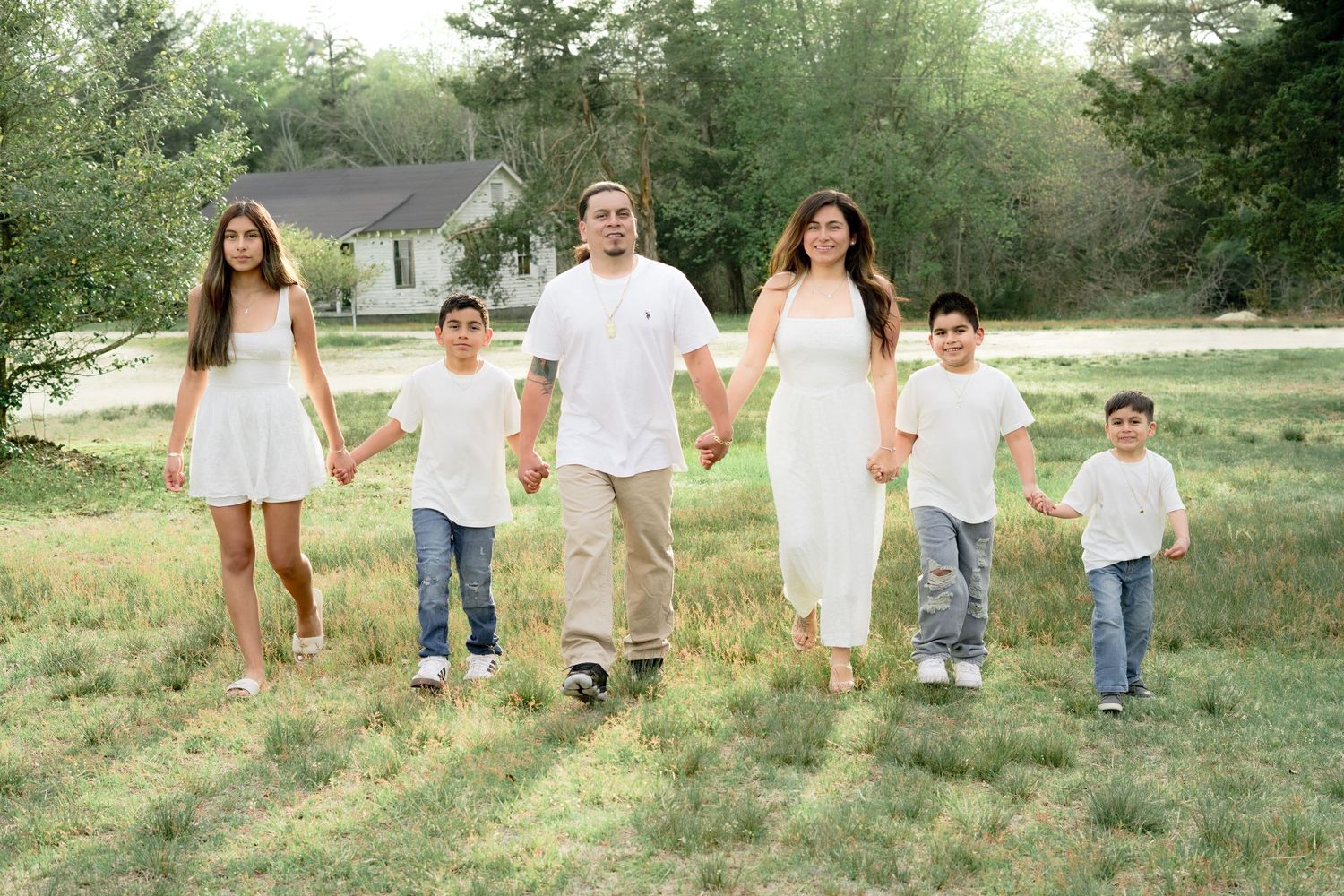 Family of five in white outfits holding hands walking through a green field near a white house.