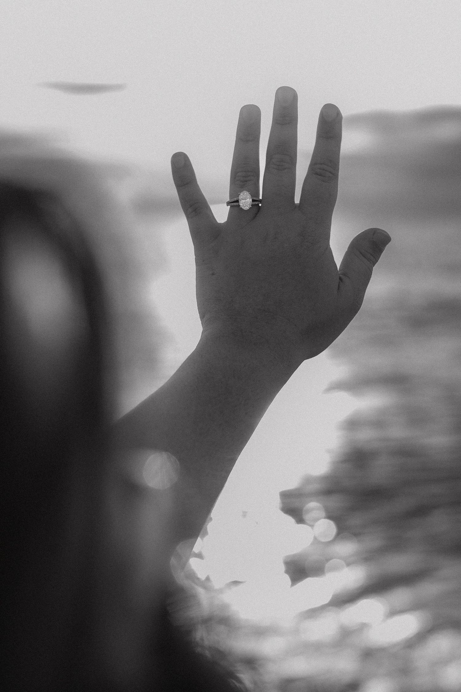 Black and white photo of a hand raised against ocean backdrop, showcasing a elegant engagement ring on finger.