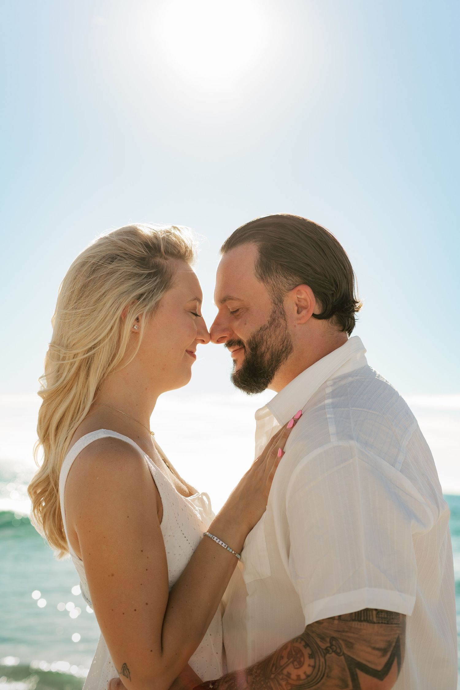 Romantic couple nearly kissing on a sunny beach, woman in white dress and tattooed man smiling face to face.