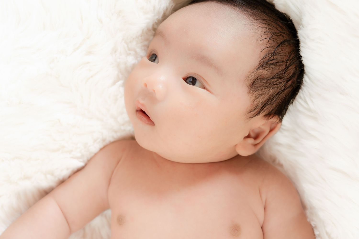 Adorable Asian newborn baby lying on soft white fluffy blanket, looking up with bright curious eyes.