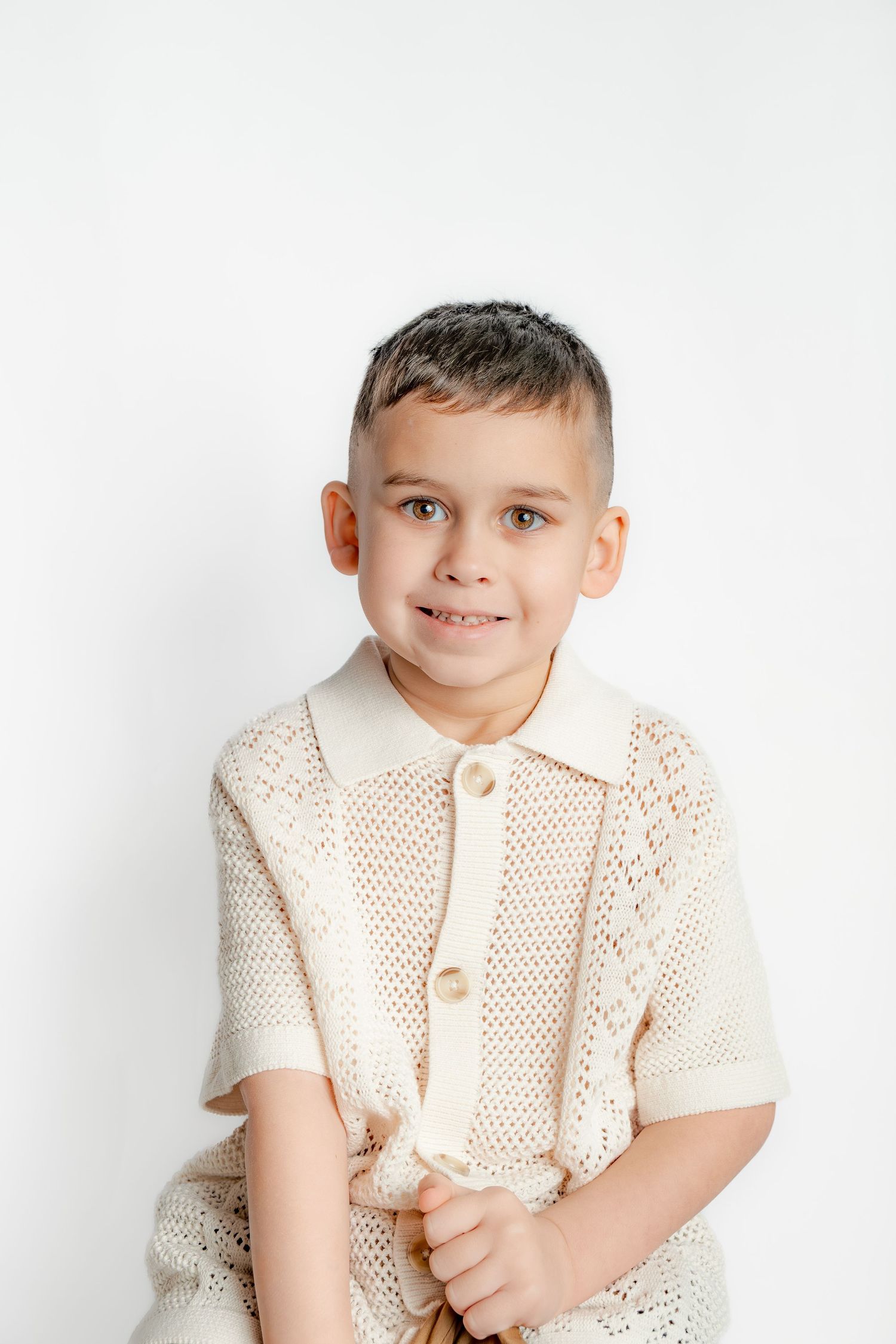 A young boy in a cream knit polo shirt smiles warmly while posing against a clean white background.