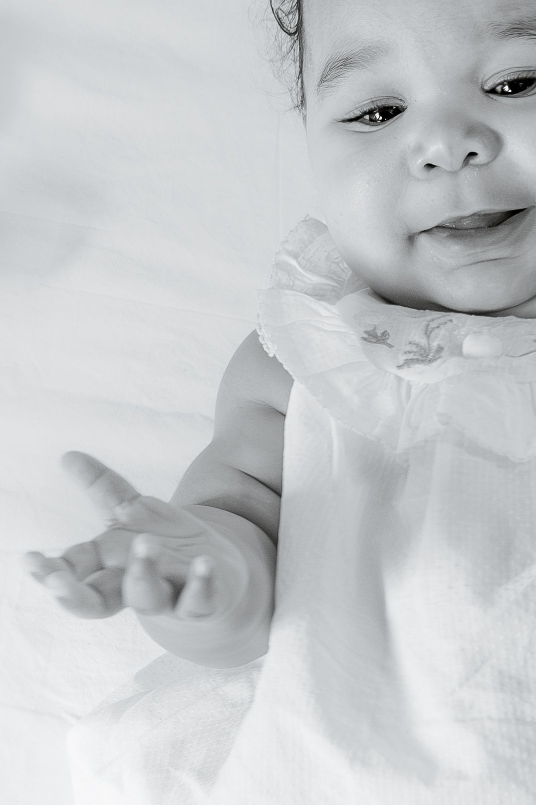 A smiling baby in a white dress reaches out her hand in a black and white portrait.