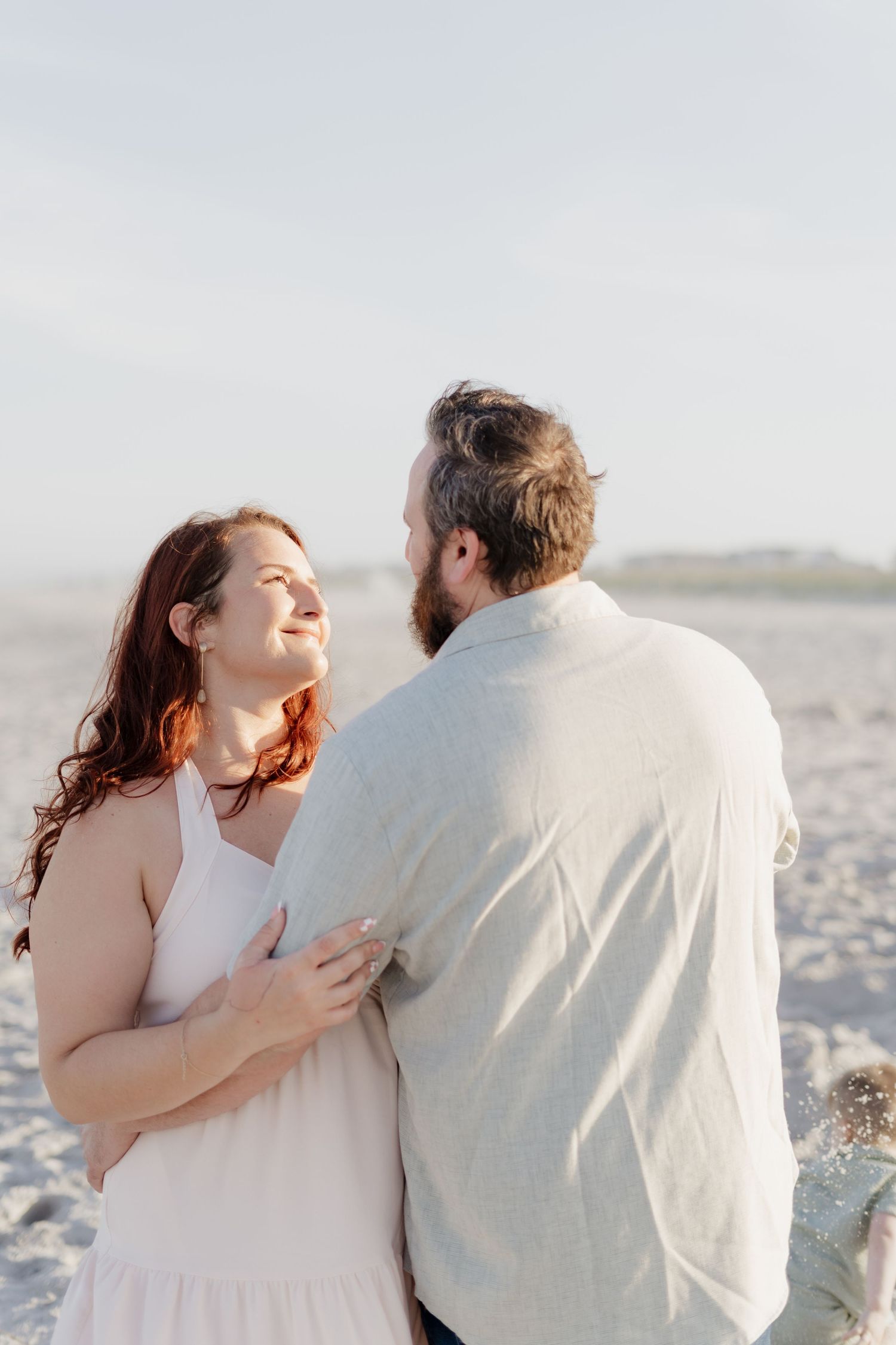 A couple embraces on a sunny beach, the woman in a white dress smiling up at her bearded partner.