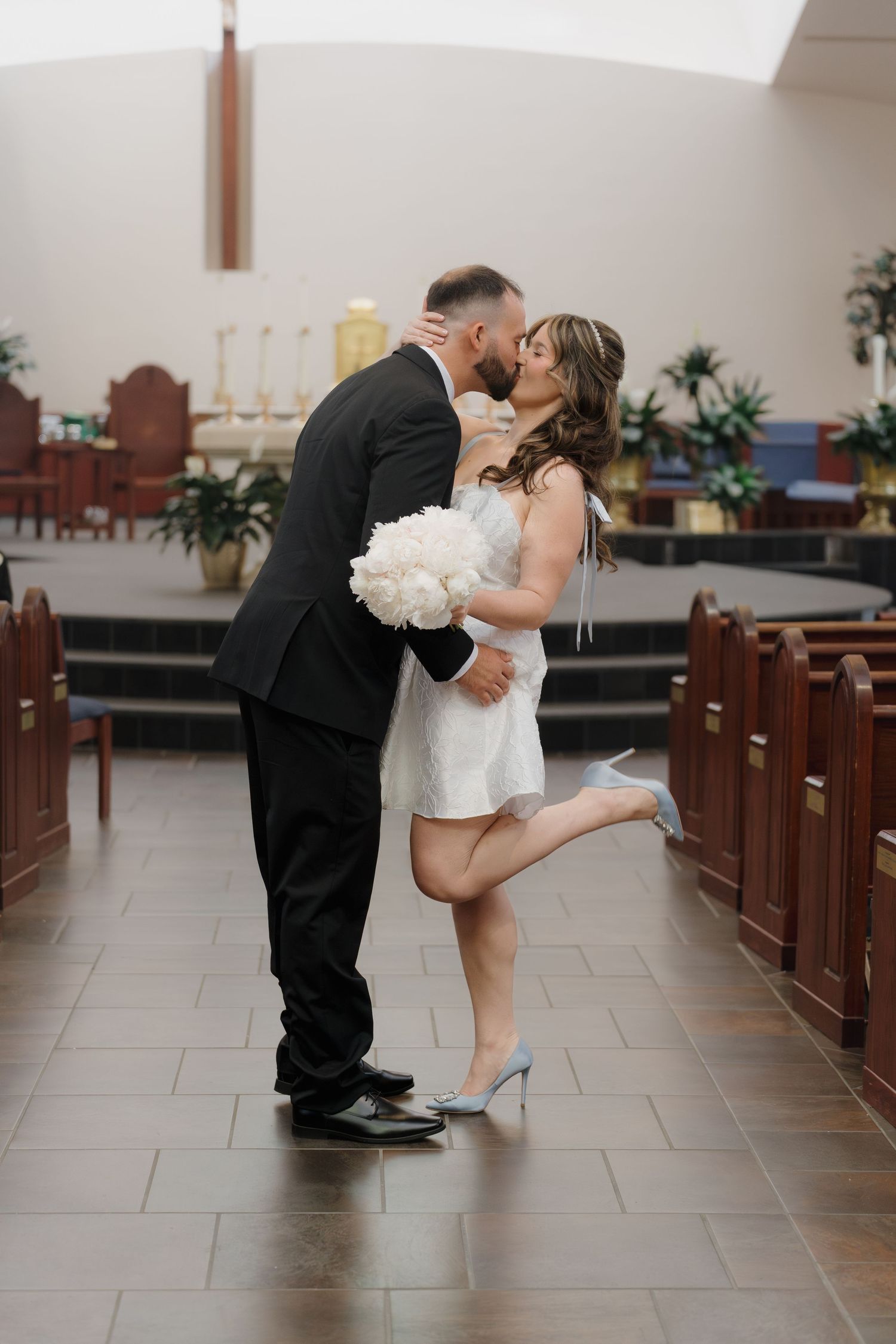 Bride and groom share a kiss in church aisle, bride in white mini dress holding bouquet, groom in black suit.